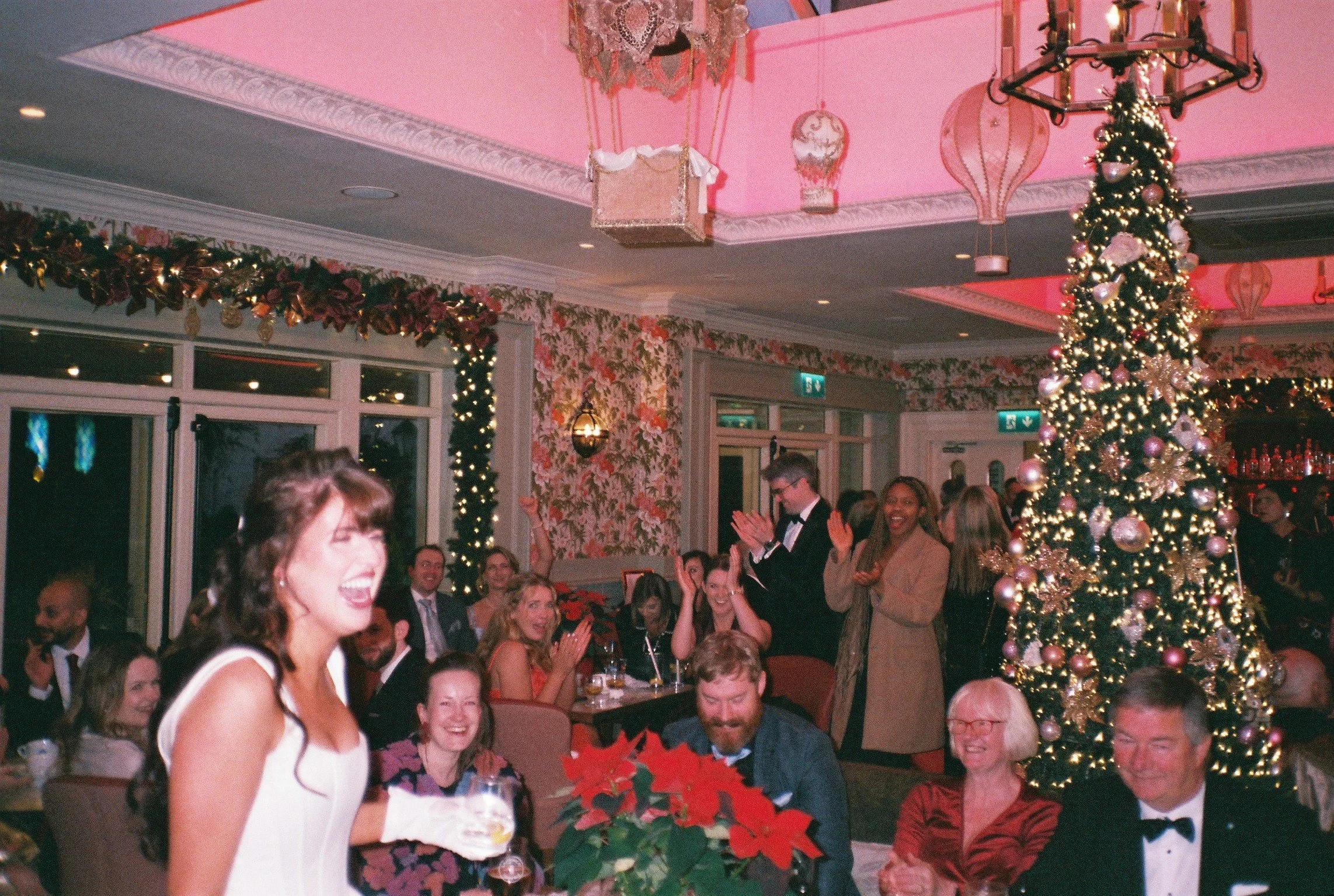 A holiday party with people celebrating around a decorated Christmas tree, with pink lighting, holiday garlands, and festive decorations.