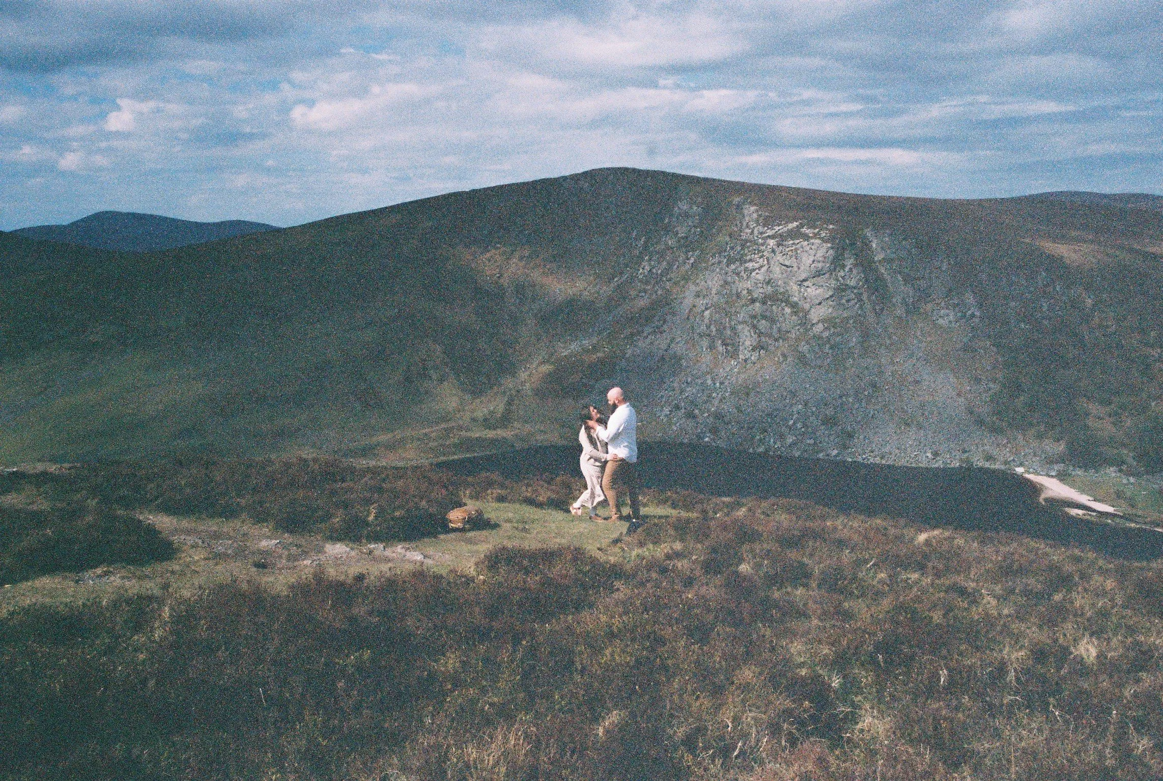 A couple stands together outdoors in a field with mountains in the background. They are close, holding hands, with the man wearing a white shirt and the woman in a light-colored dress. The sky is partly cloudy.