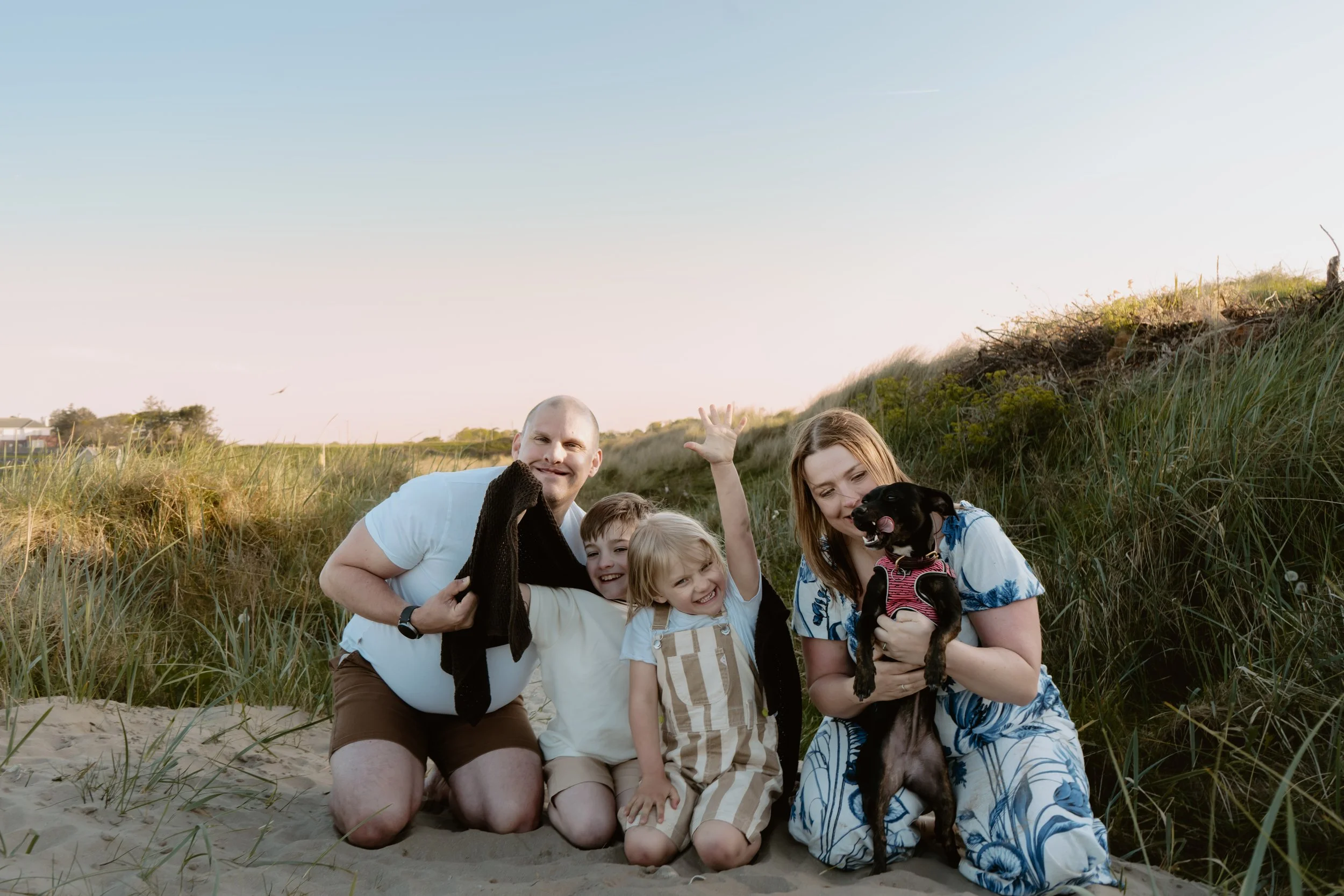 Family of four with a dog enjoying time on the beach in the early evening, surrounded by sand and tall grass.