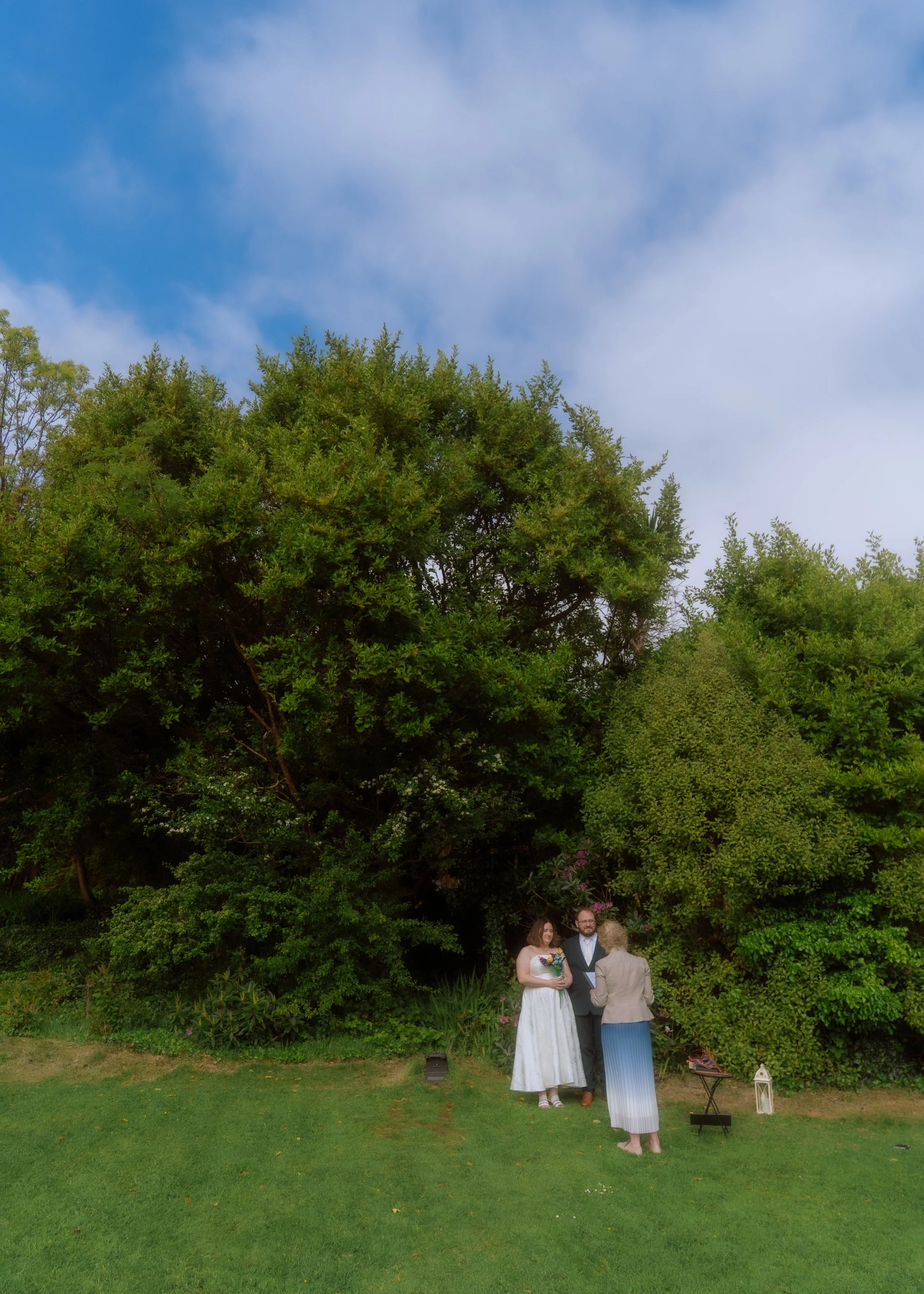A wedding ceremony taking place outdoors in a garden with green grass, trees, and a partly cloudy sky. Four people are present: a bride in a white dress holding a bouquet, an officiant in a dark suit, a man in a gray suit, and a woman in a light-colored jacket and long skirt. There is a small table with decorations and a lantern on the grass.