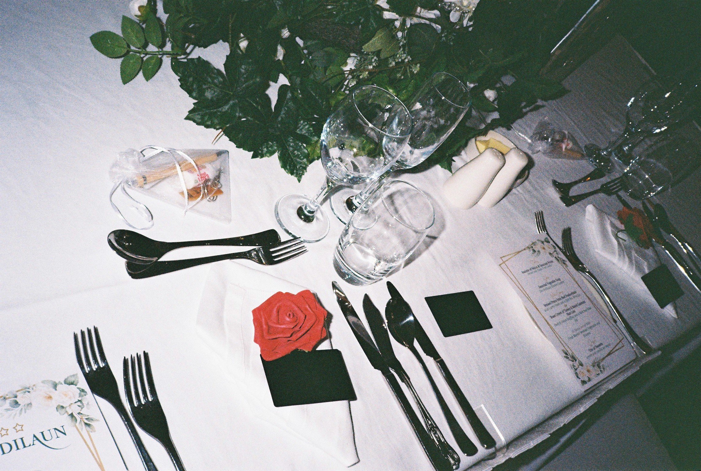 Table set for a celebration with black napkins, silverware, wine glasses, a red rose napkin, and a menu card. There is a green plant in the background.