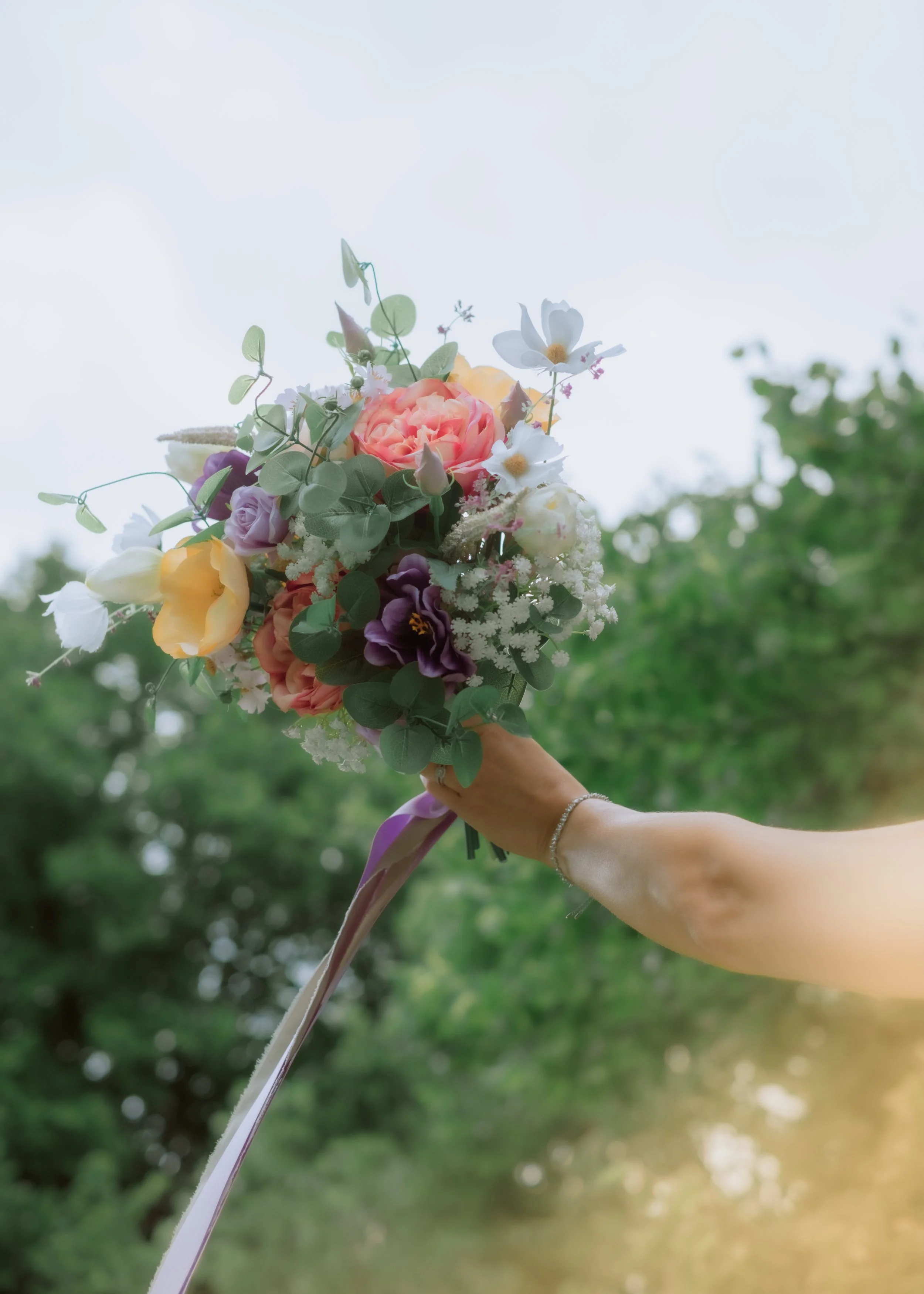 Hand holding a colorful bouquet of flowers against a blurred outdoor green background.