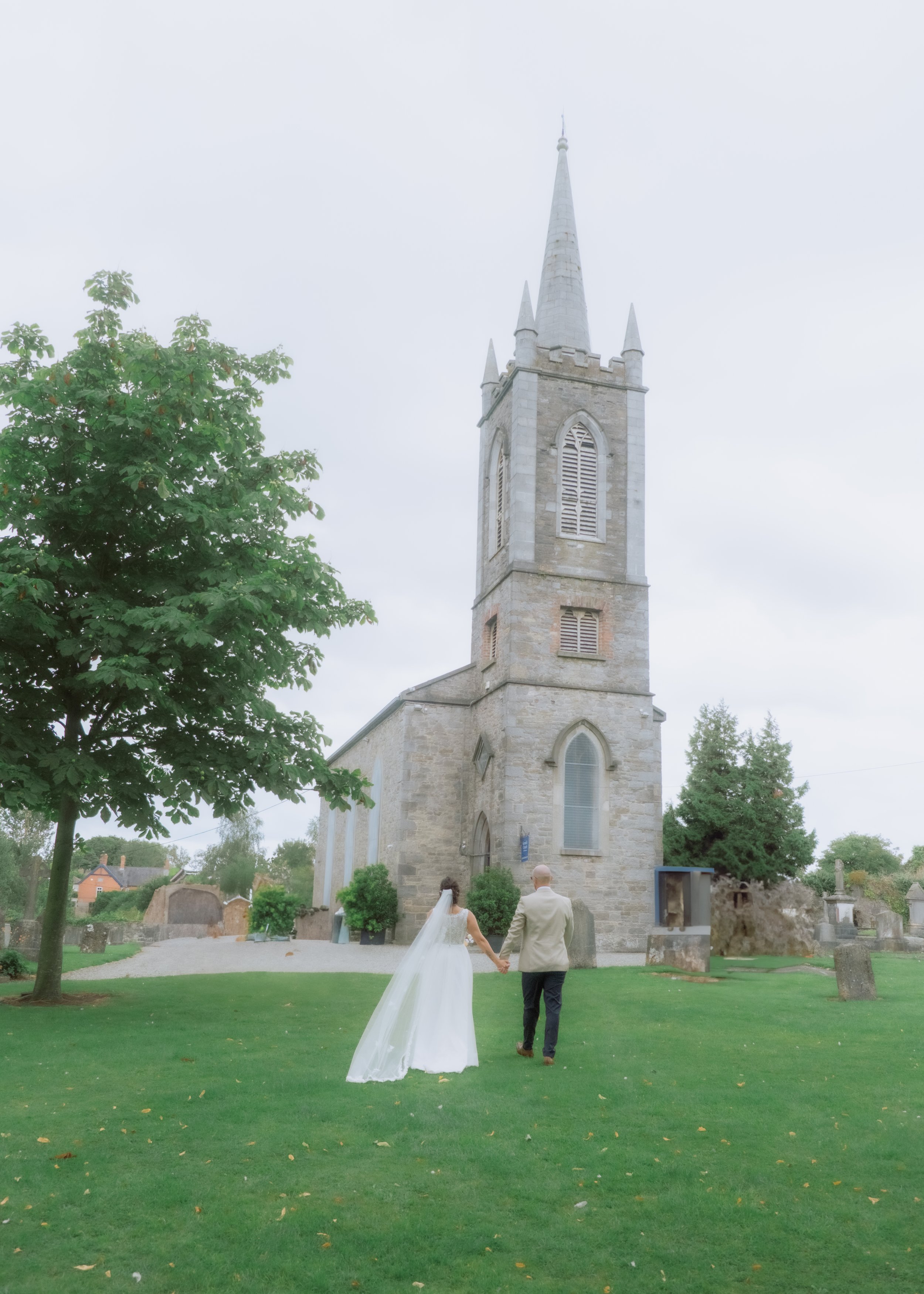 A bride and groom holding hands walking on a green lawn in front of a stone church with a tall steeple, surrounded by trees and gravestones.