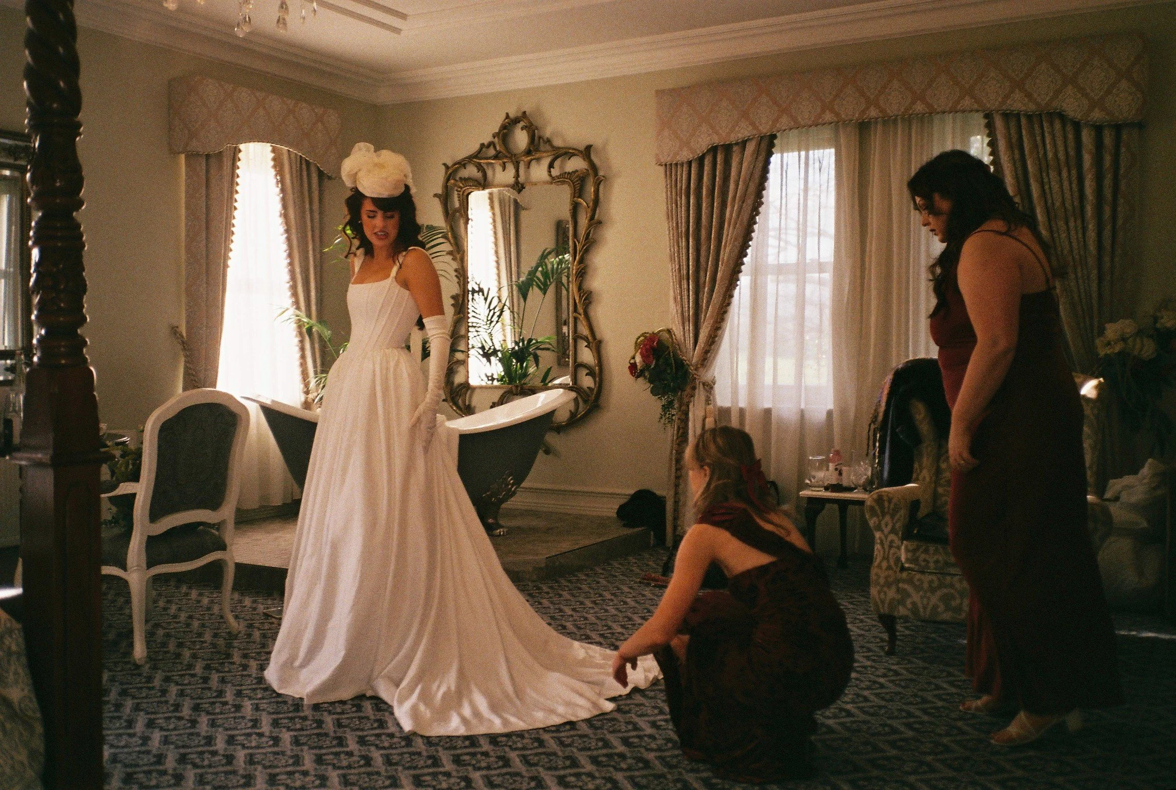 A woman in a white dress and long gloves is standing in a room, possibly preparing for a special event. Another woman in a dark dress is nearby, and a girl is sitting on the floor, looking up. The room has elegant curtains, a large mirror with an orn