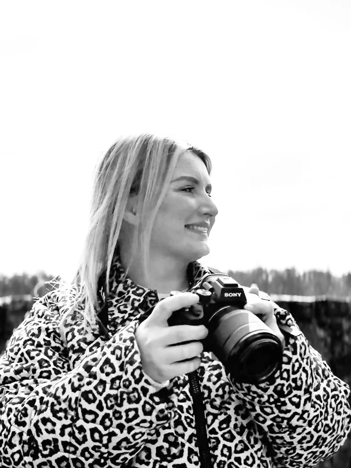 A woman with blonde hair wearing a leopard print jacket, holding a Sony camera outdoors, smiling.