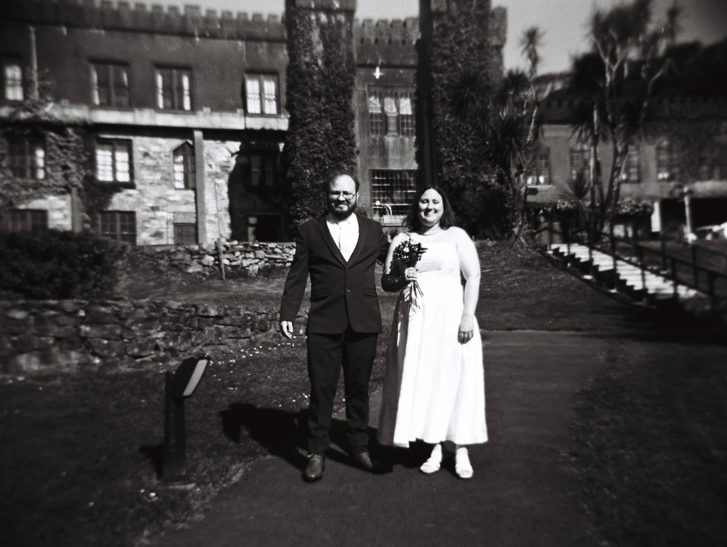 A black and white photo of a couple dressed in wedding attire, standing outdoors in front of a stone building with multiple windows and a tall tree, with a small stone wall and stairs nearby.