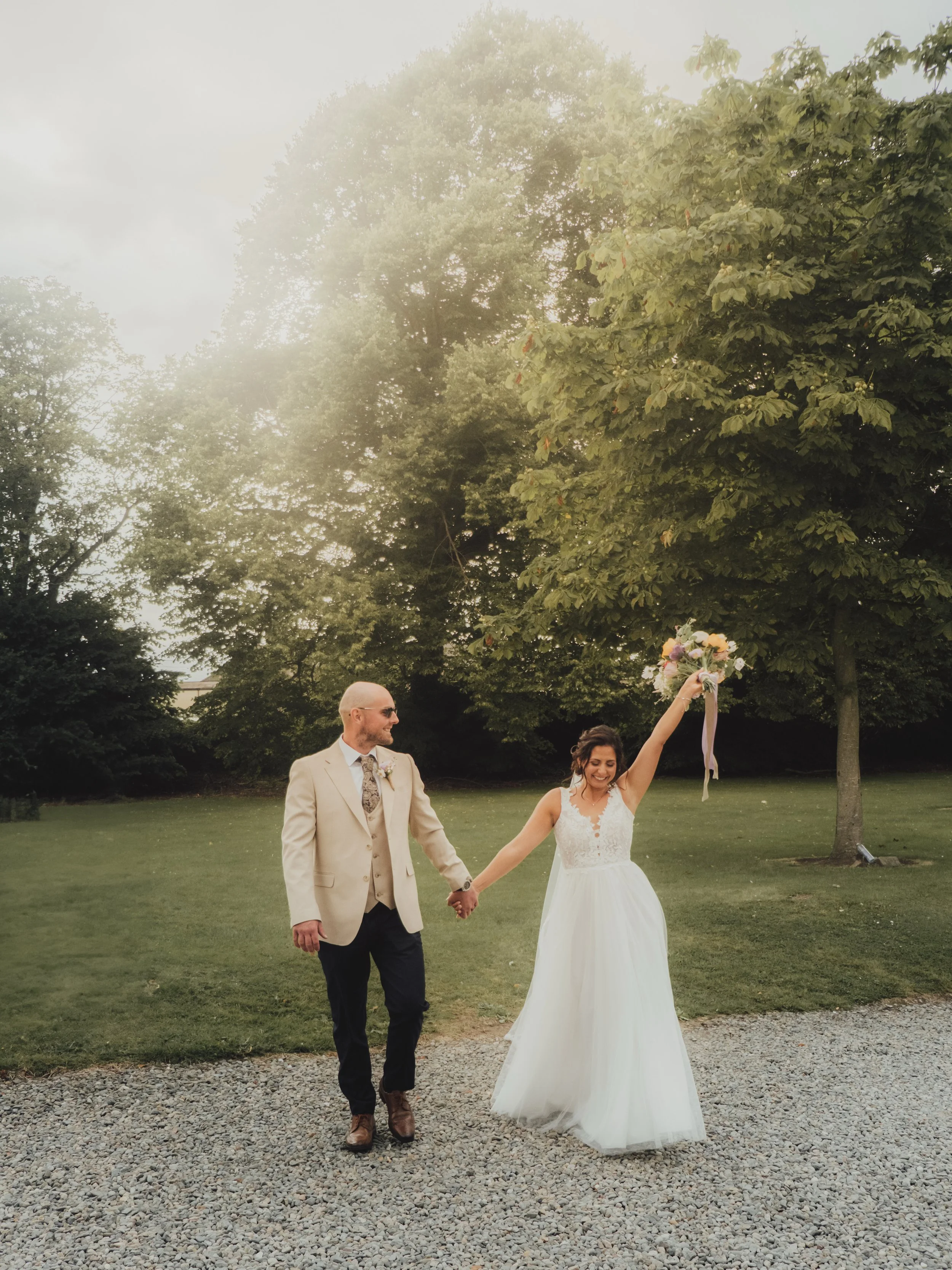 A bride and groom walk hand-in-hand outdoors on a gravel path, the bride holding a bouquet and smiling, with trees in the background.