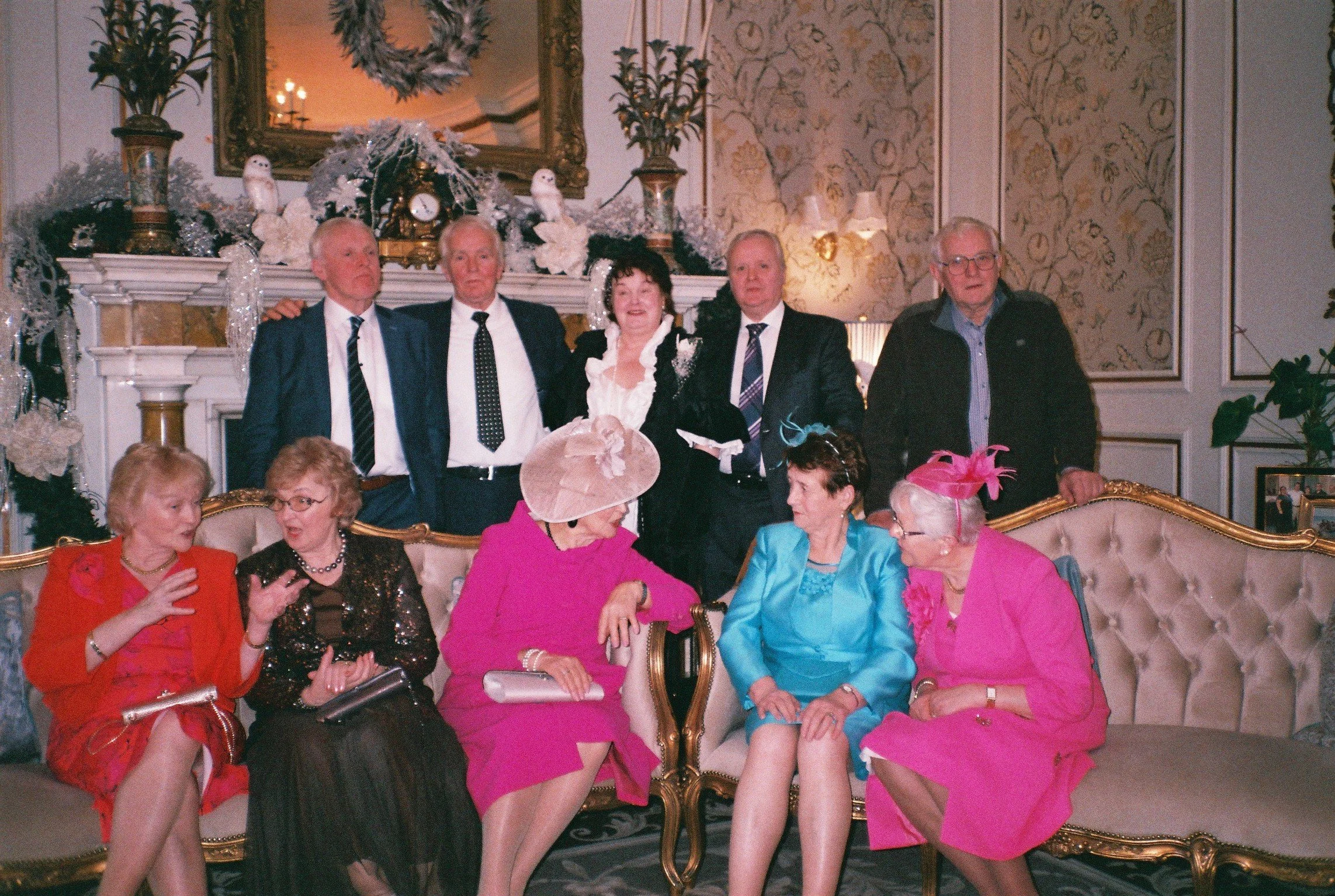 Group of elderly women and men gathered in a decorated room for a social event. Five women are seated in the foreground, dressed in colorful and formal clothing, with some wearing hats. Several men are standing behind them, dressed in suits and ties,