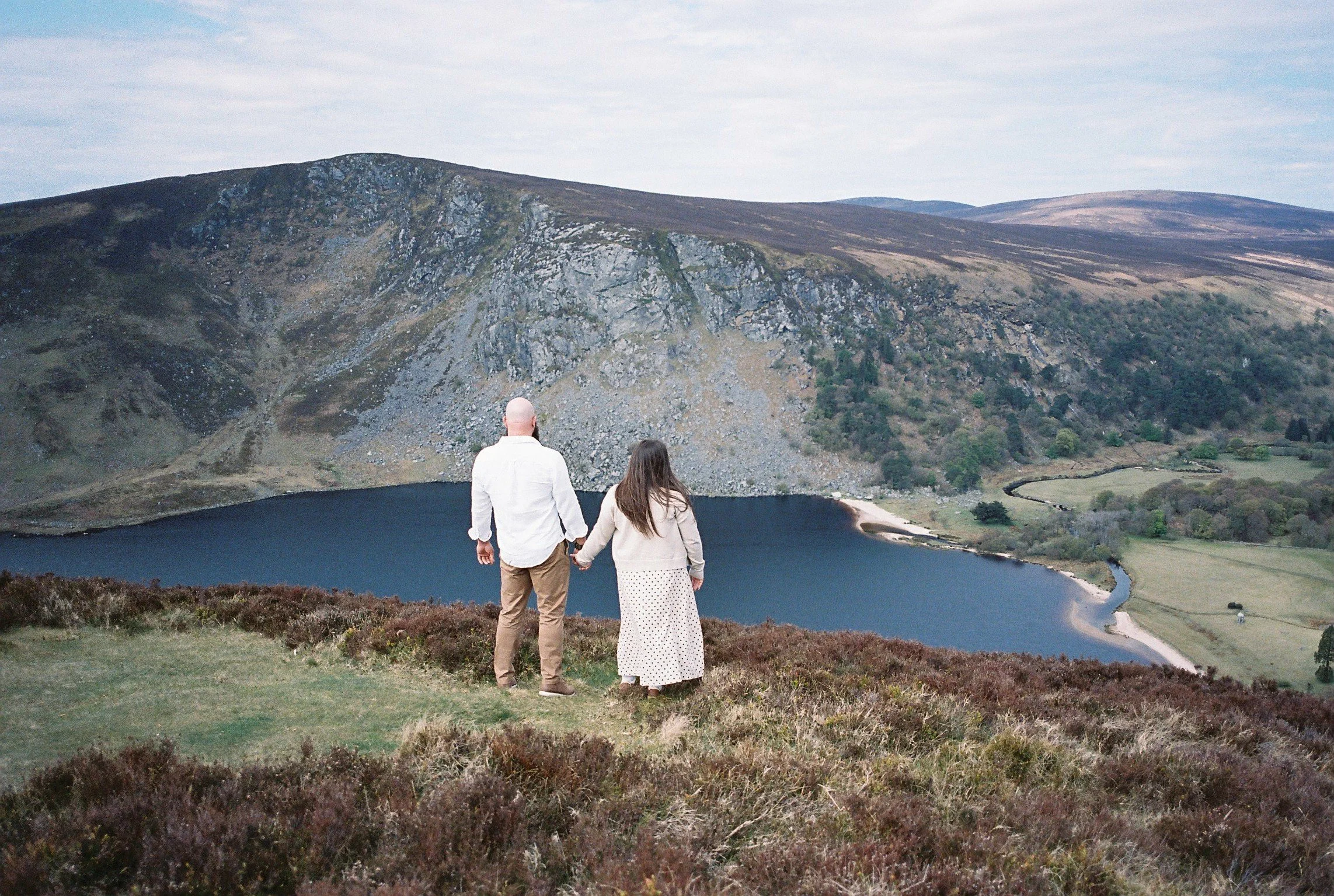 A couple holding hands and facing a mountain lake surrounded by hills and grassy landscape.