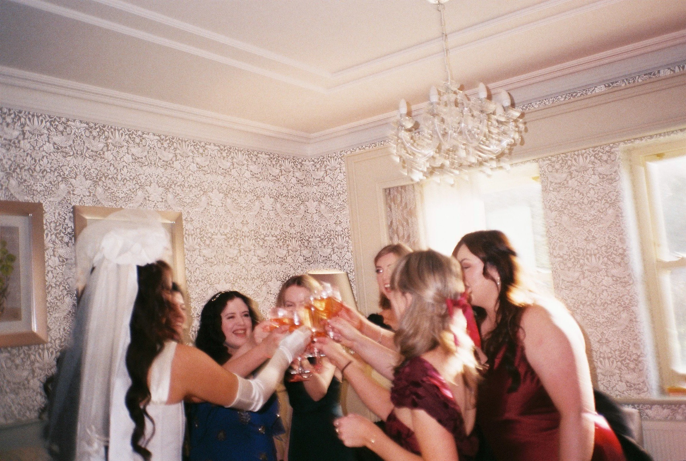 Group of women at a celebration, exchanging bouquets of flowers in a decorated room with ornate wallpaper and a chandelier.