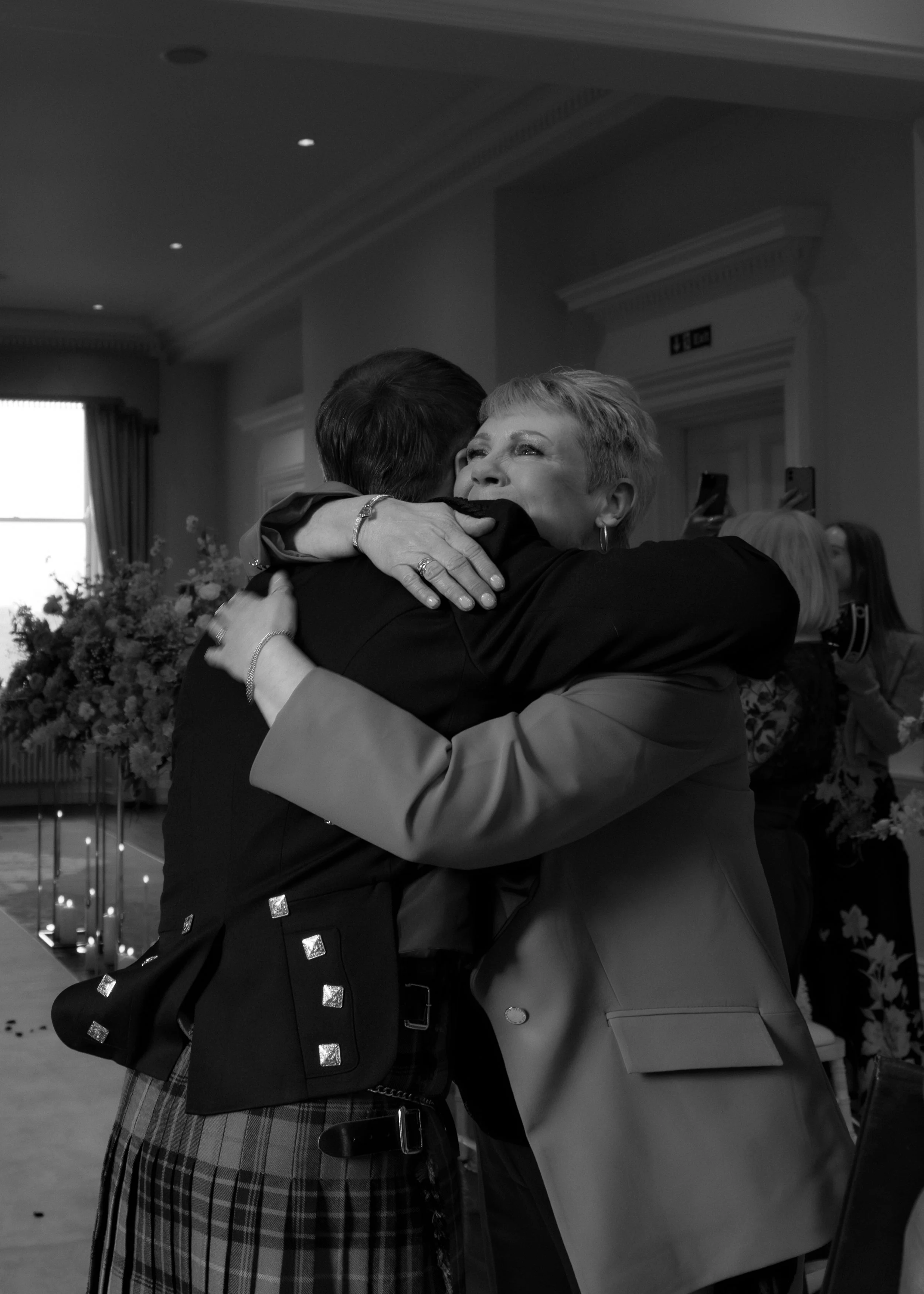 Two women hugging warmly, one appears older, in a formal setting with floral arrangements and guests in the background.
