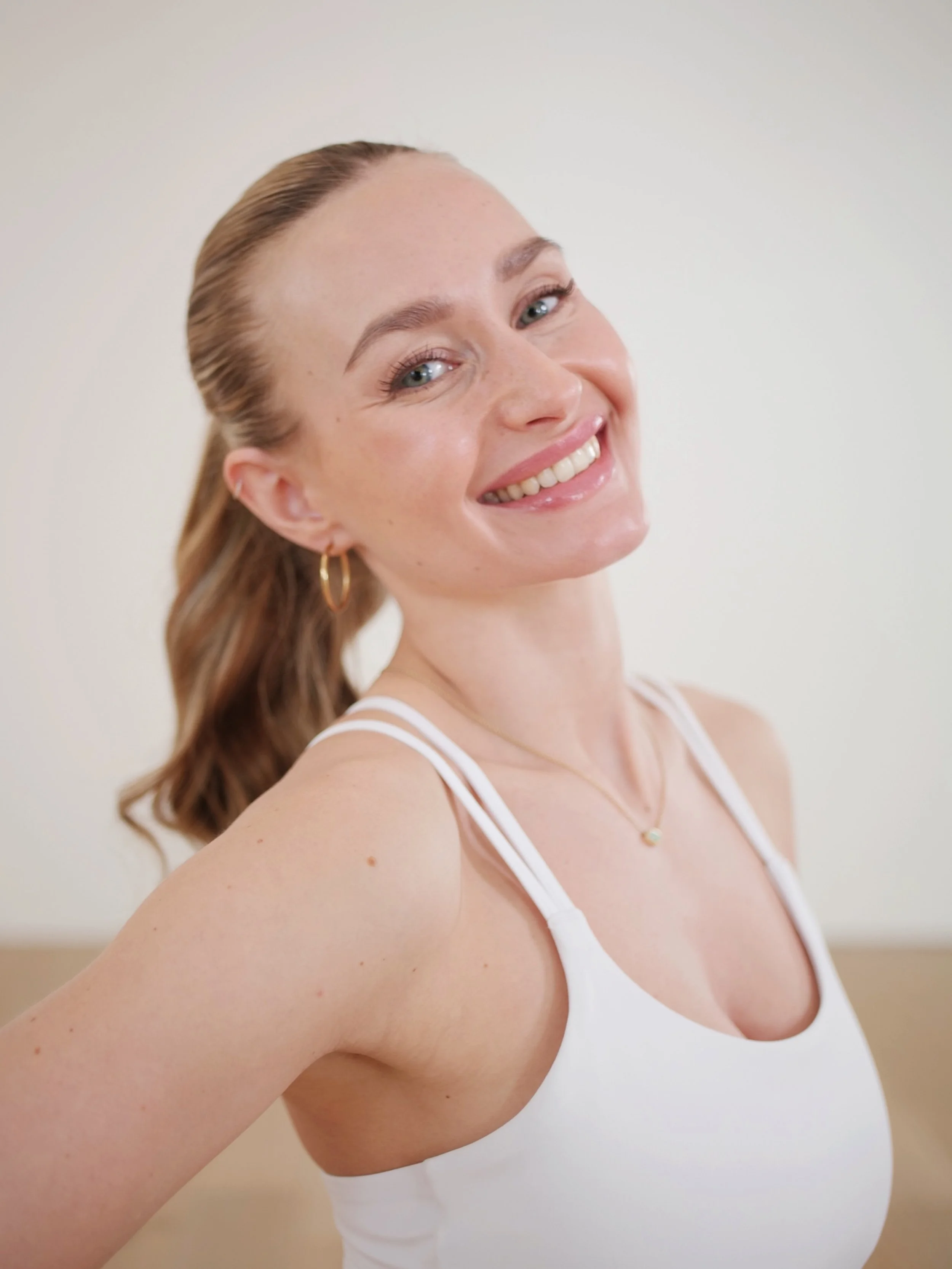 Nahaufnahme eines lächelnden Mädchens mit hellen Haaren, das Arm und Schulter zeigt, trägt eine weiße Top und kleine goldene Ohrringe gegen einen neutralen Hintergrund.