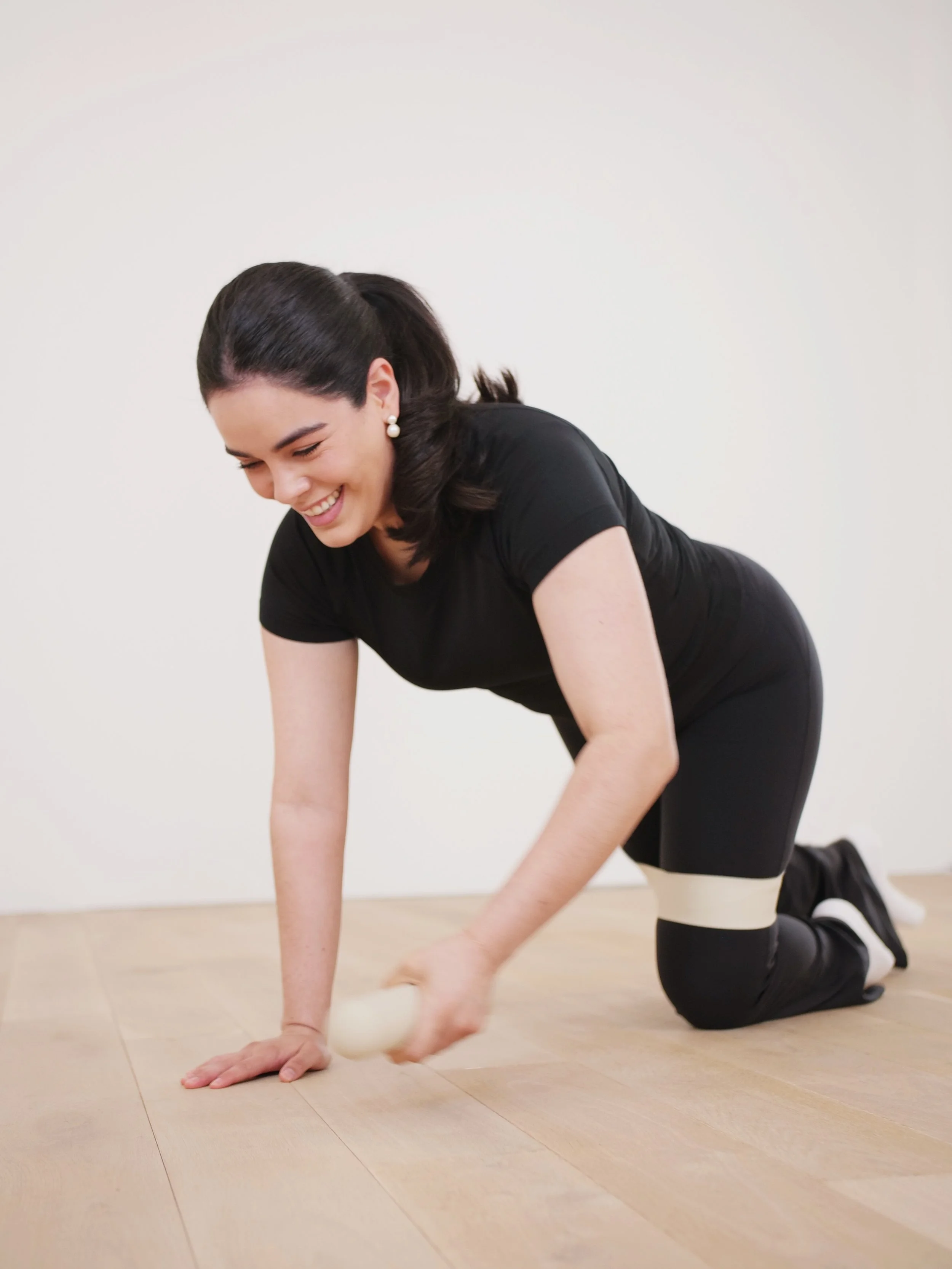 Eine junge Frau in schwarzen Kleid mit weißen Strümpfen kniet auf einem Holzboden, macht Yoga und lächelt.