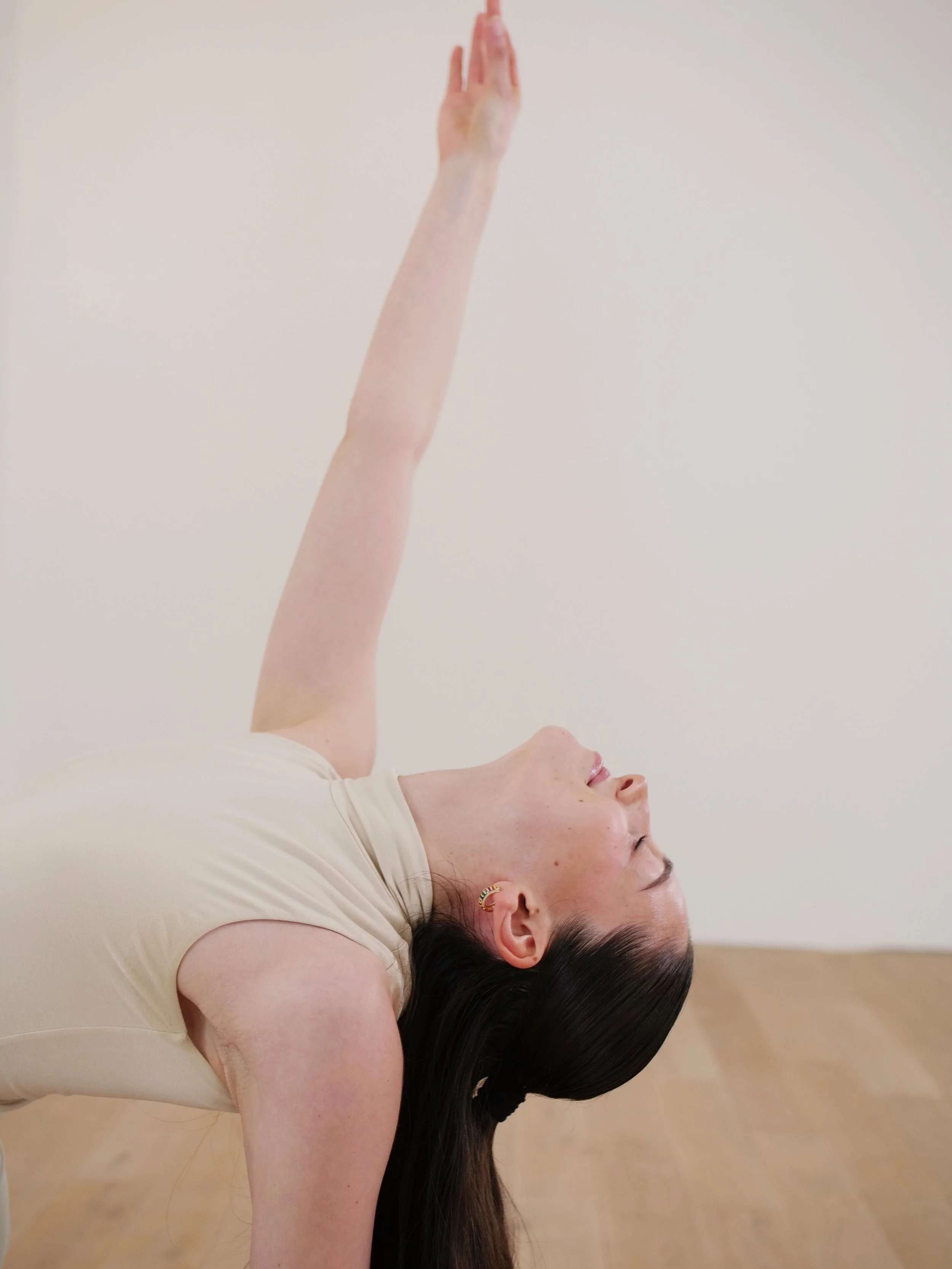 Frau liegt auf dem Boden, macht eine Yoga-Pose mit nach oben gestreckten Armen und gebeugtem Oberkörper, mit langem dunklem Haar und Ohrringen, in einem hellen Raum.