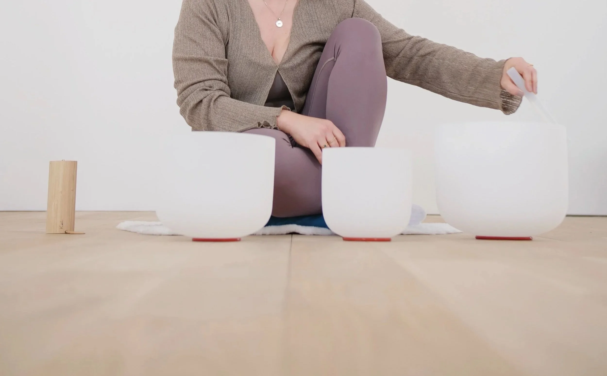 Eine Person sitzt auf dem Boden, umgeben von weißen Klangschalen und einem kleinen Holzblock, in einem Raum mit hellem Hintergrund.
