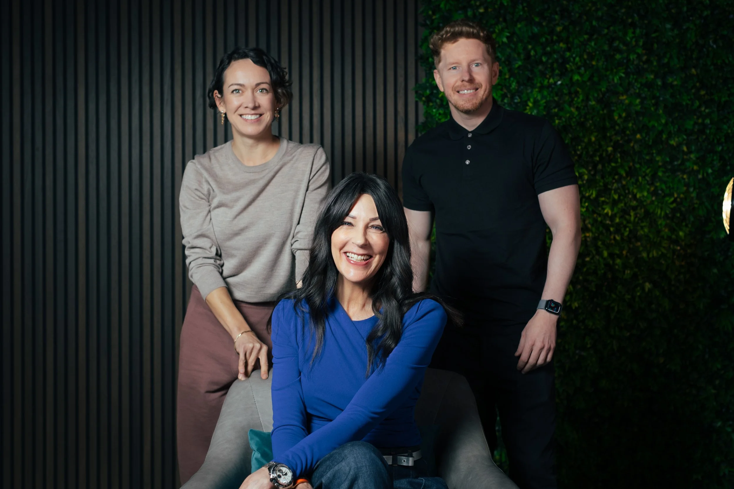 Group of four diverse people smiling, with a woman seated in front and three standing behind her, against a patterned wall and greenery.