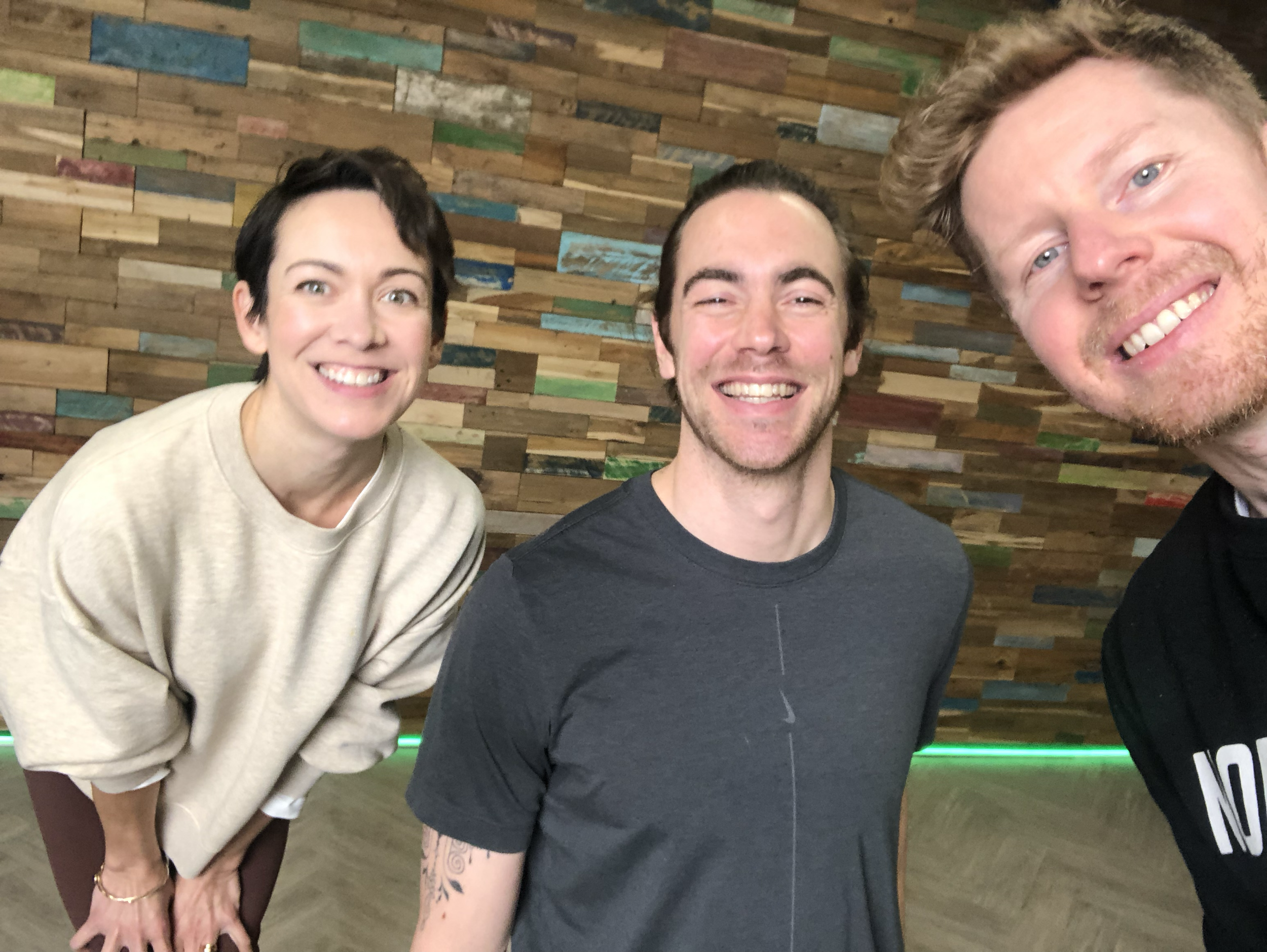 A group of three smiling friends taking a selfie indoors against a wooden wall with multicolored planks