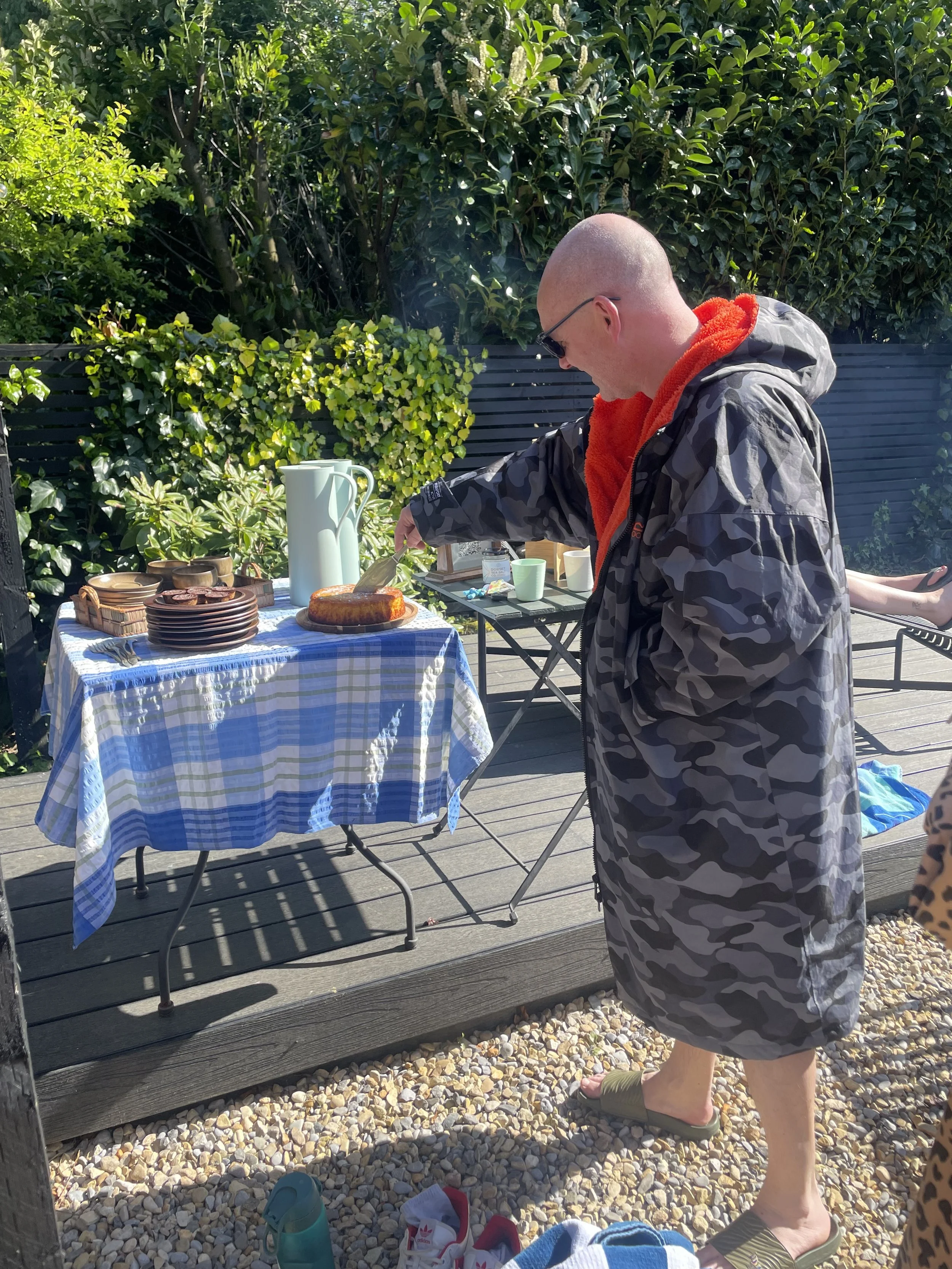 A person with a shaved head wearing sunglasses, a camouflage jacket, and green slides, standing outdoors on a wooden deck next to a table with baked goods and a large white thermos, with green foliage in the background.