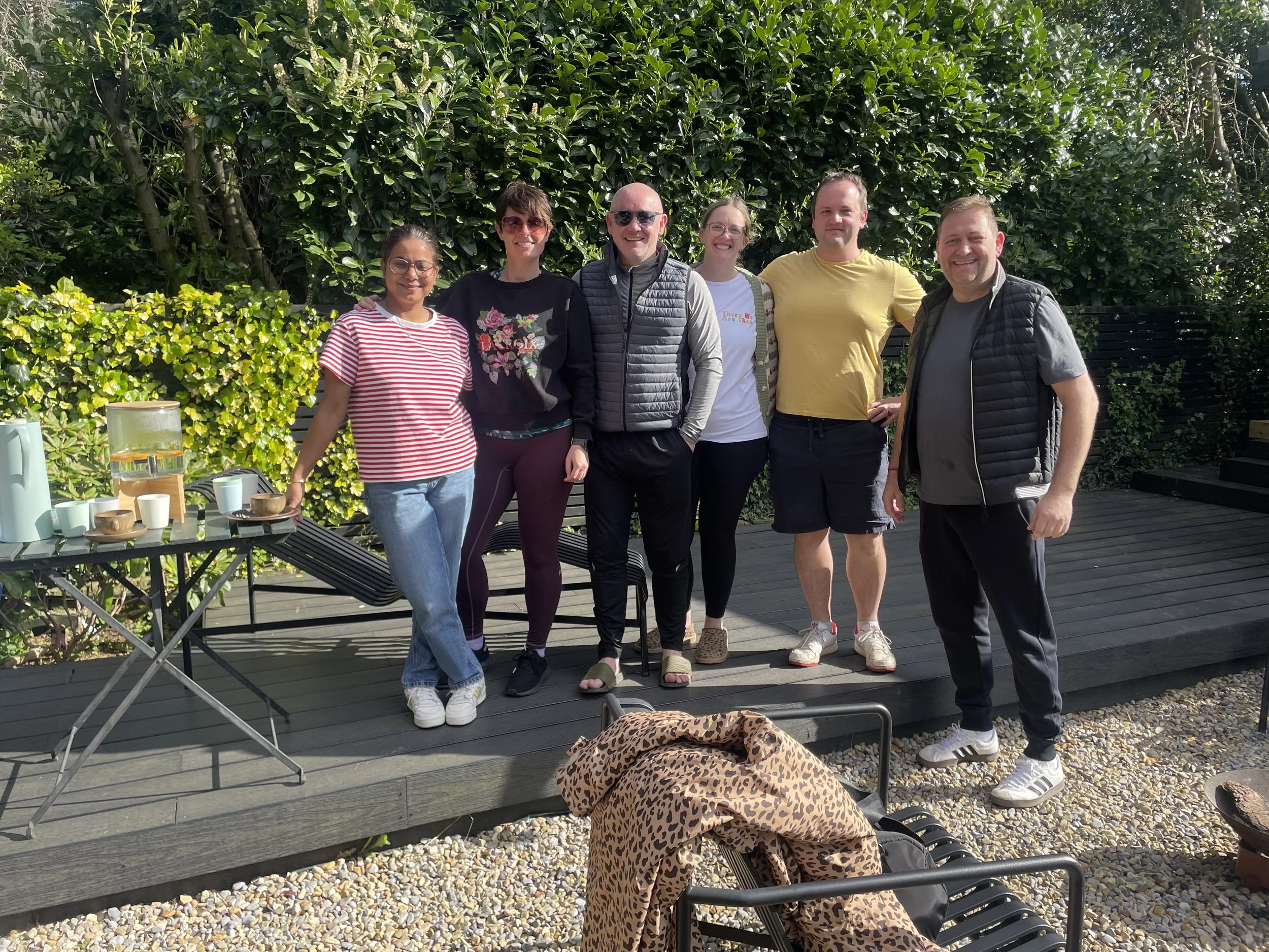 Group of seven smiling people standing outdoors on a deck in front of lush green bushes, with a table holding various cups and a leopard-print bag in the foreground.