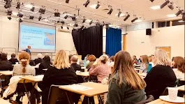 People attending a presentation or seminar in a classroom or conference room, facing a speaker and a projection screen.
