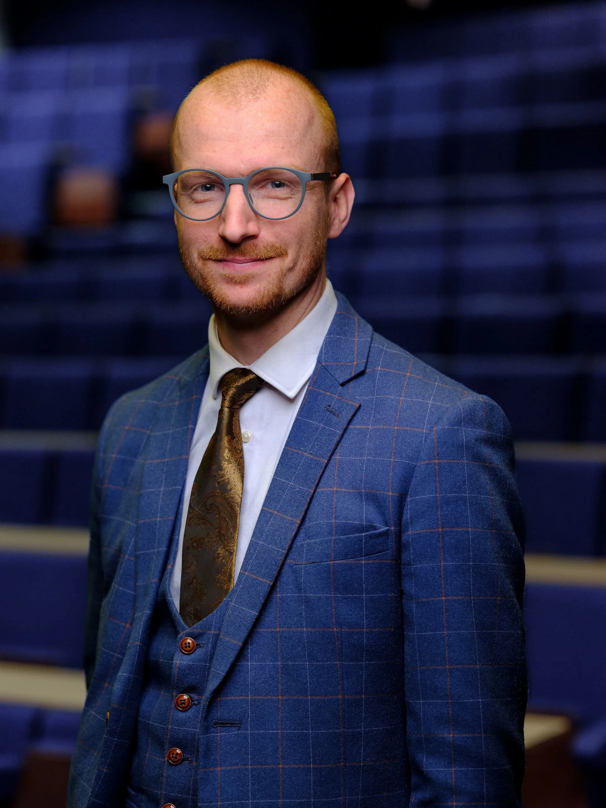 Portrait of a man with glasses, a beard, and a mustache, wearing a blue checkered suit, white shirt, and a brown patterned tie, standing in front of a blurred blue background.
