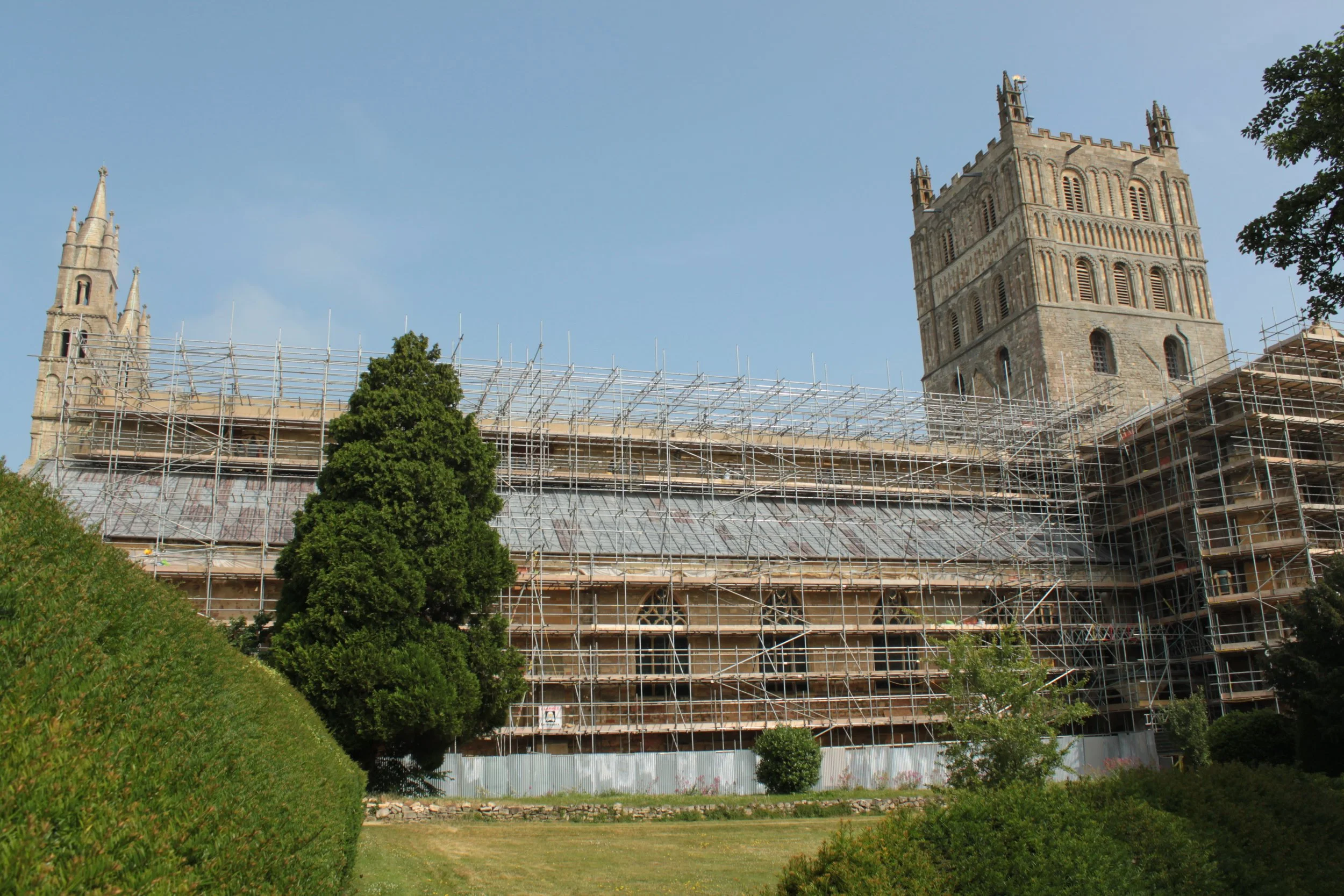 Scaffolding surrounding a historic stone building during renovation, with a grassy foreground and clear sky.