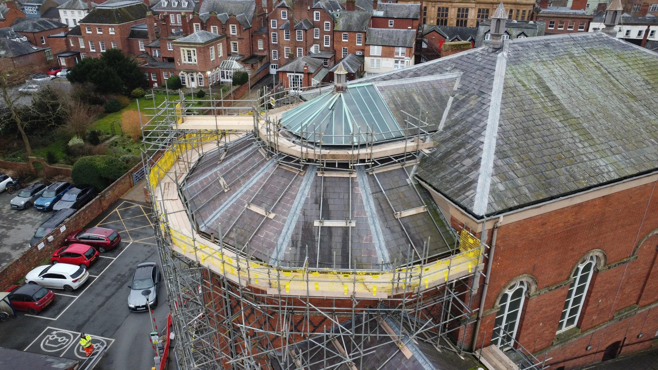 Aerial view of building under renovation with scaffolding around a domed section and surrounding residential area, with parked cars.