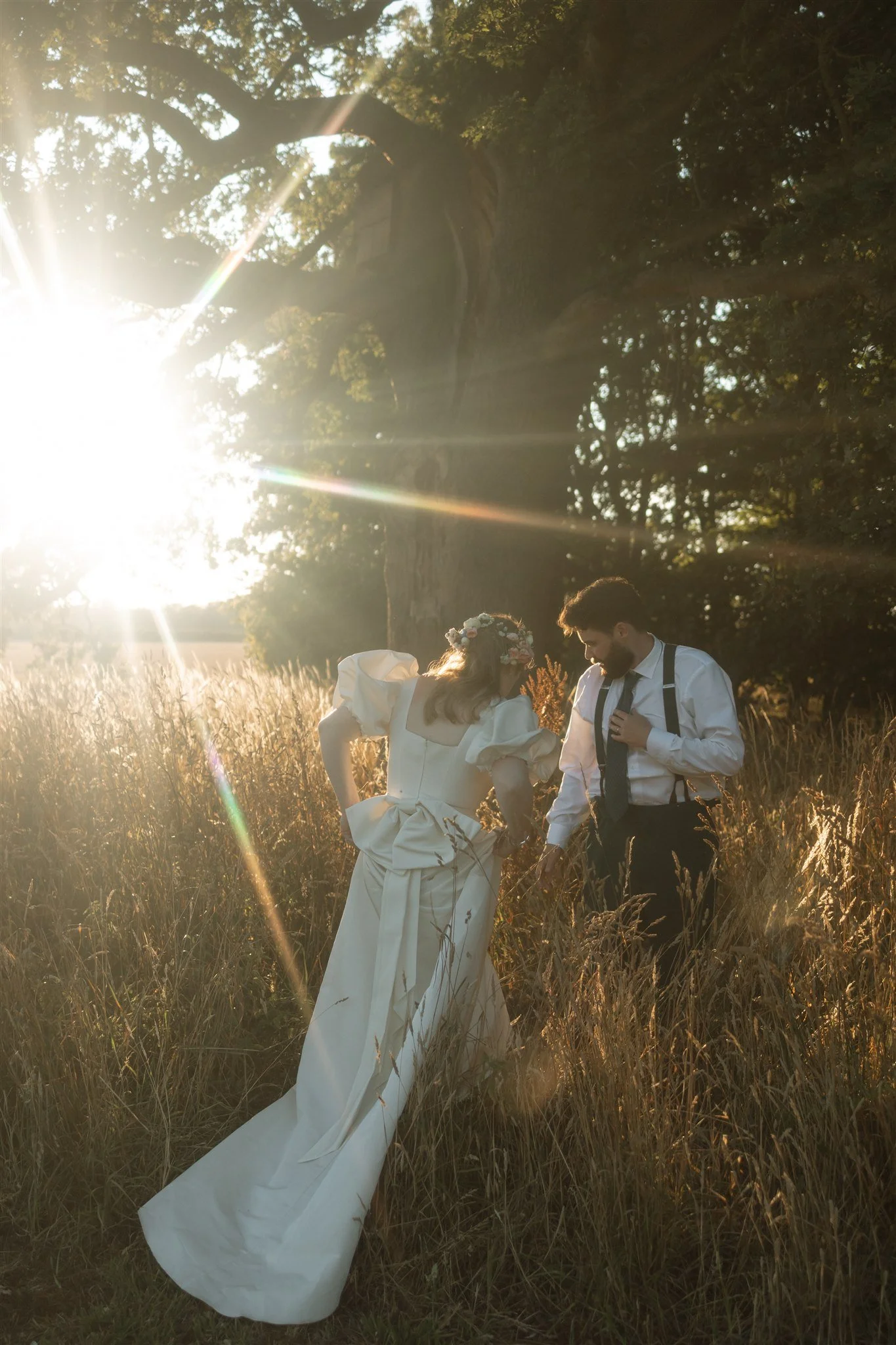 A bride and groom walking through tall grass in a sunlit field during sunset, with the bride wearing a white wedding gown and the groom in a white shirt with suspenders.