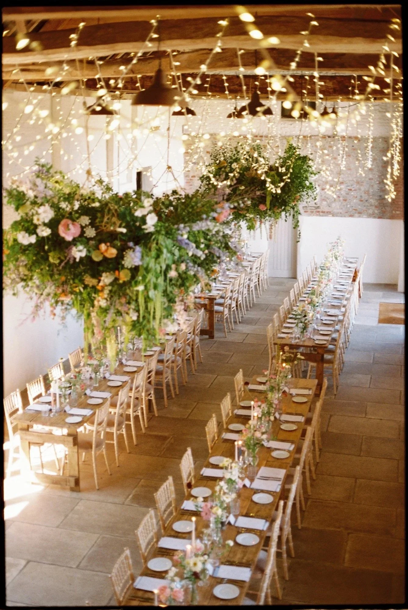 A decorated banquet hall with long wooden tables, floral centerpieces, string lights, and hanging pendant lamps.