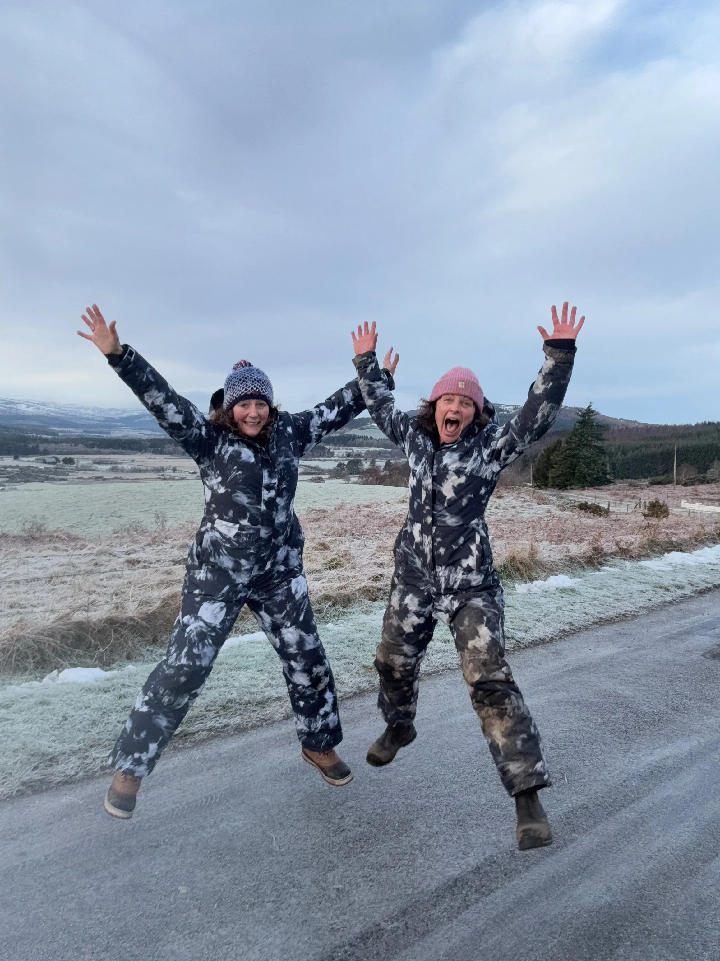 Two women in camouflage winter outfits jumping in the air with arms raised, standing on the side of a frosty road in a rural landscape, with snow-capped mountains and cloudy sky in the background.