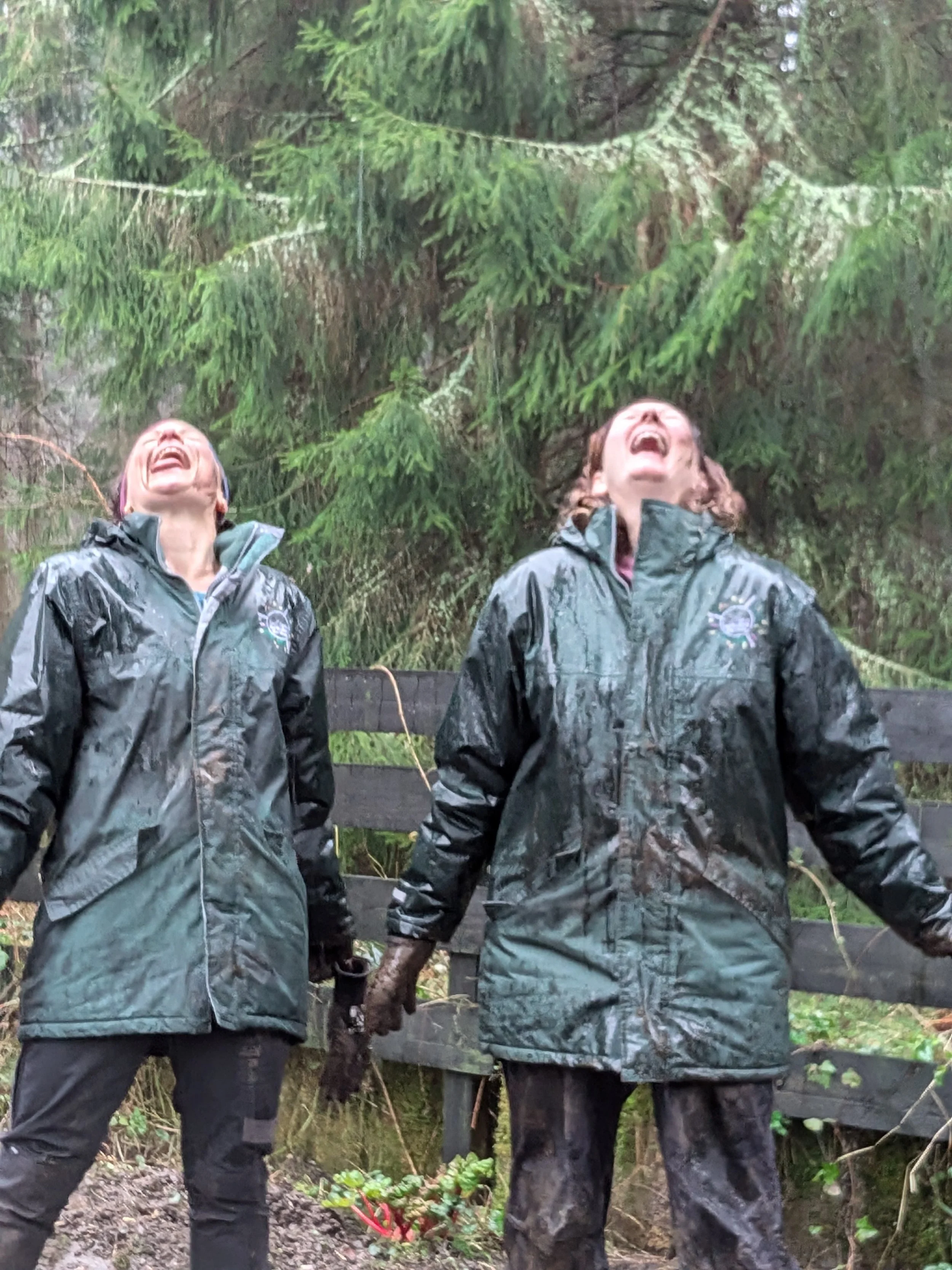 Two women in waterproof jackets are laughing, standing outdoors with lush green trees and a wooden fence in the background.