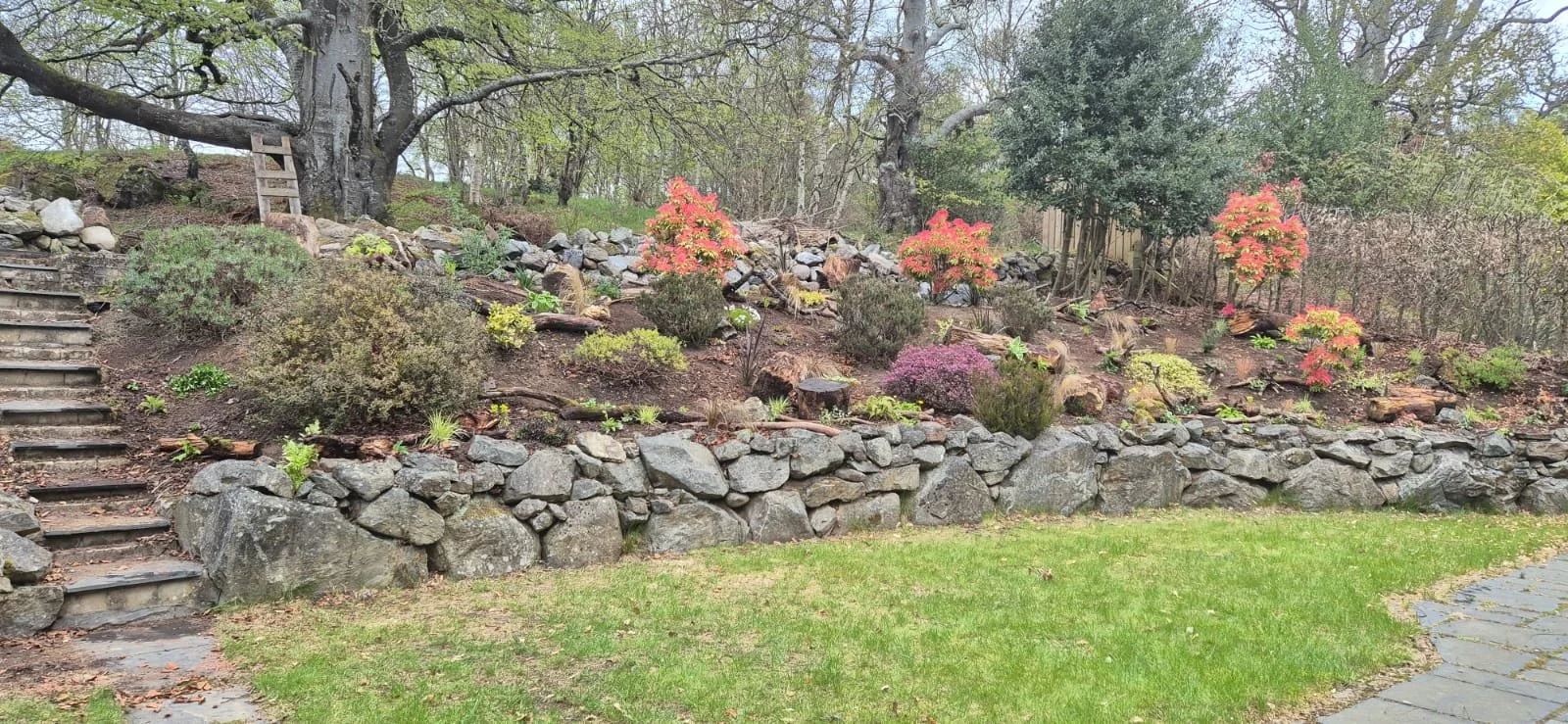 A person planting flowers in a garden bed surrounded by rocks, with gardening tools and equipment nearby.