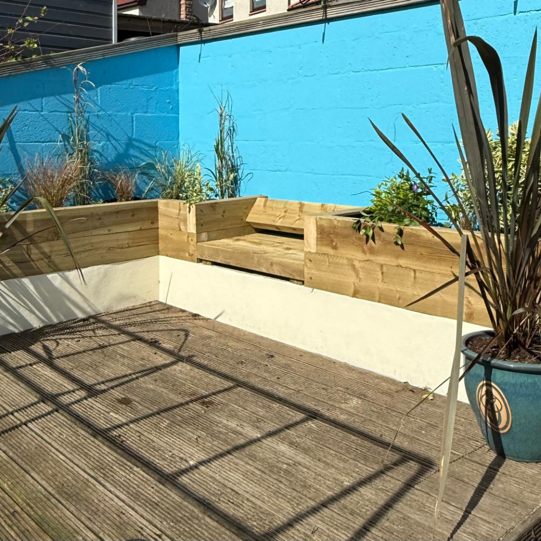 Small outdoor patio with wooden decking, a wooden raised garden bed with plants, potted plants, and a bright blue wall in the background.