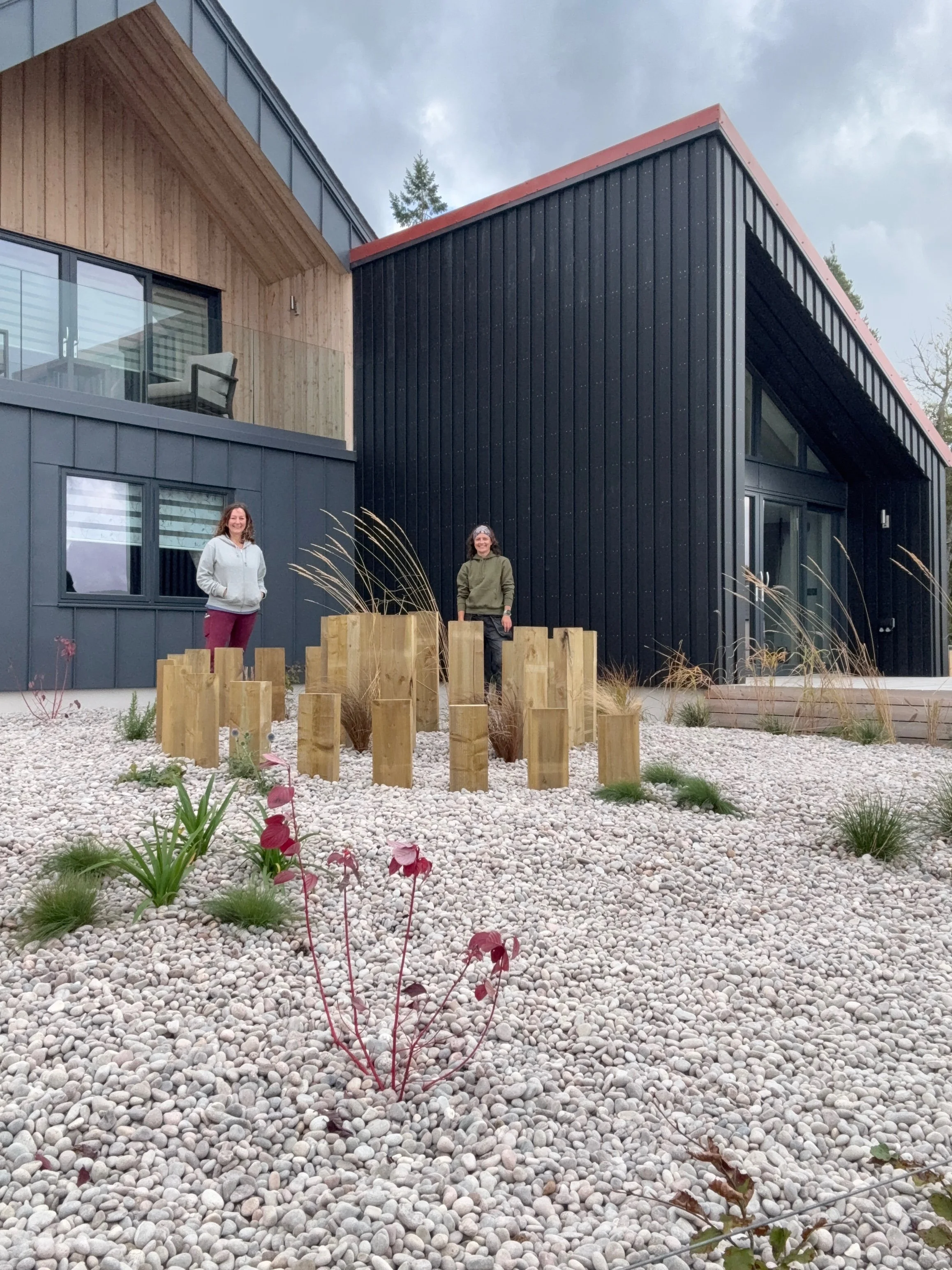 Two women stand in front of a modern black and wood-sided house with a gravel yard and wooden landscaping features.
