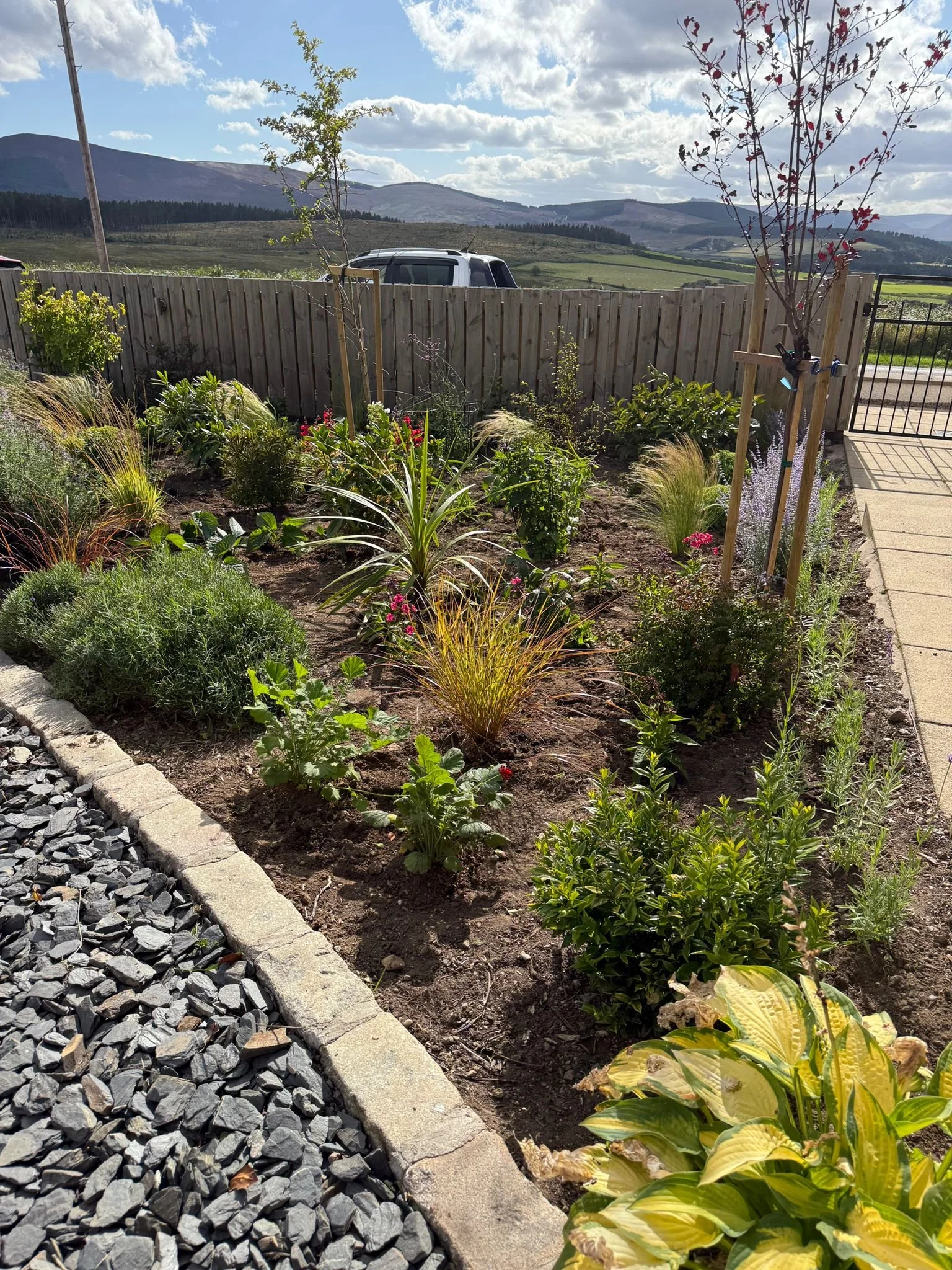A garden with various plants and flowers, bordered by a stone edge and gravel on the left. A wooden fence separates the garden from the scenic outdoor background with mountains, open fields, and a partly cloudy sky.