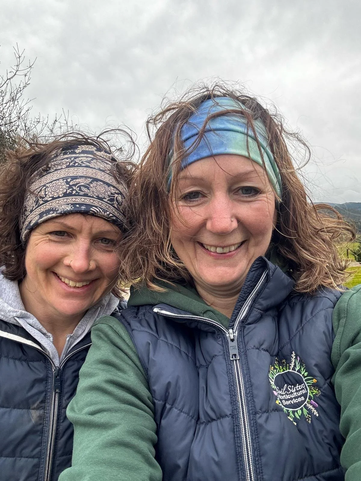 Two women smiling outdoors on a cloudy day, wearing headbands and matching navy blue vests with green sleeves, with a natural landscape in the background.