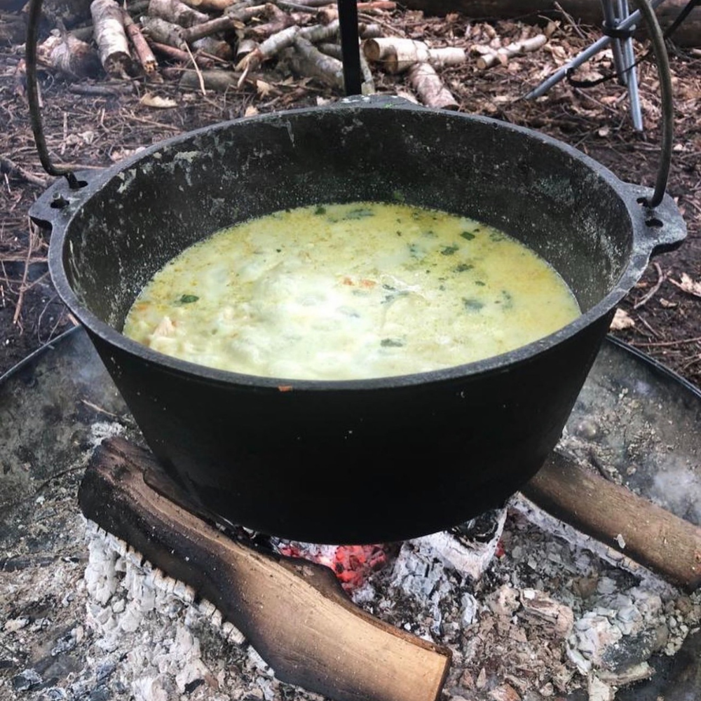A large black cast iron pot with soup or stew, simmering over an open campfire on logs, in an outdoor setting with dirt and sticks in the background.