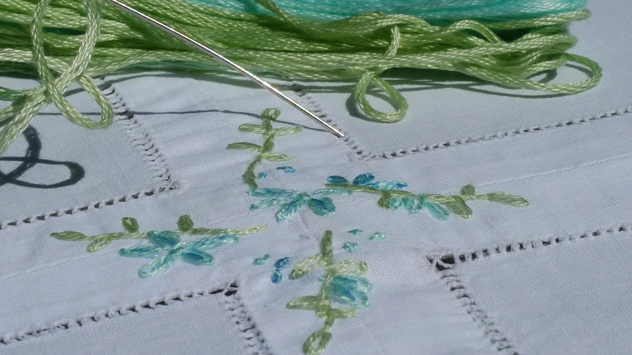 Various shades of green embroidery floss and threads arranged on a beige fabric with some embroidered green patterns.