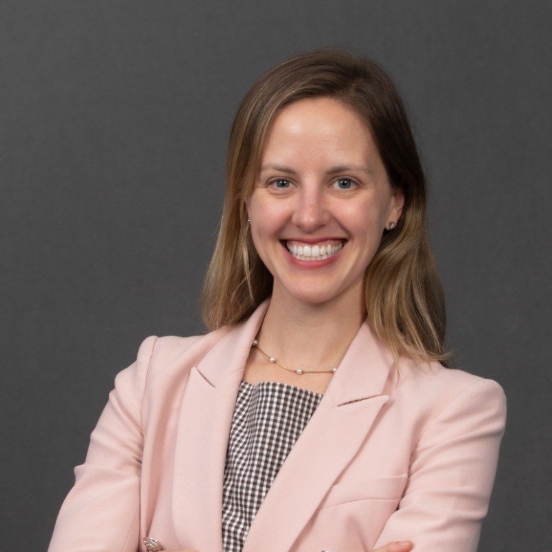 A woman with shoulder-length light brown hair smiling, wearing a pink blazer over a black and white checkered top, standing against a dark grey background.