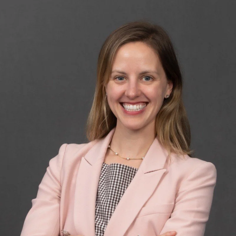 A woman with shoulder-length light brown hair, smiling, wearing a light pink blazer over a black and white checkered top, and a delicate necklace, standing against a gray background.