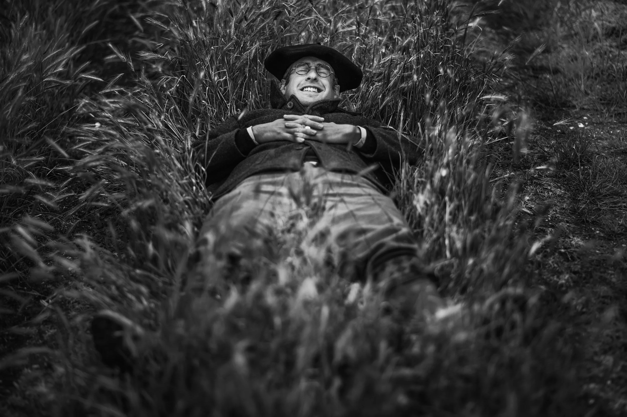 Armand Heitz, a French winemaker lying on his back in tall grass, smiling with eyes closed, wearing glasses, a hat, and a coat. Vines Chassagne Montrachet 1er Cru Maltroie