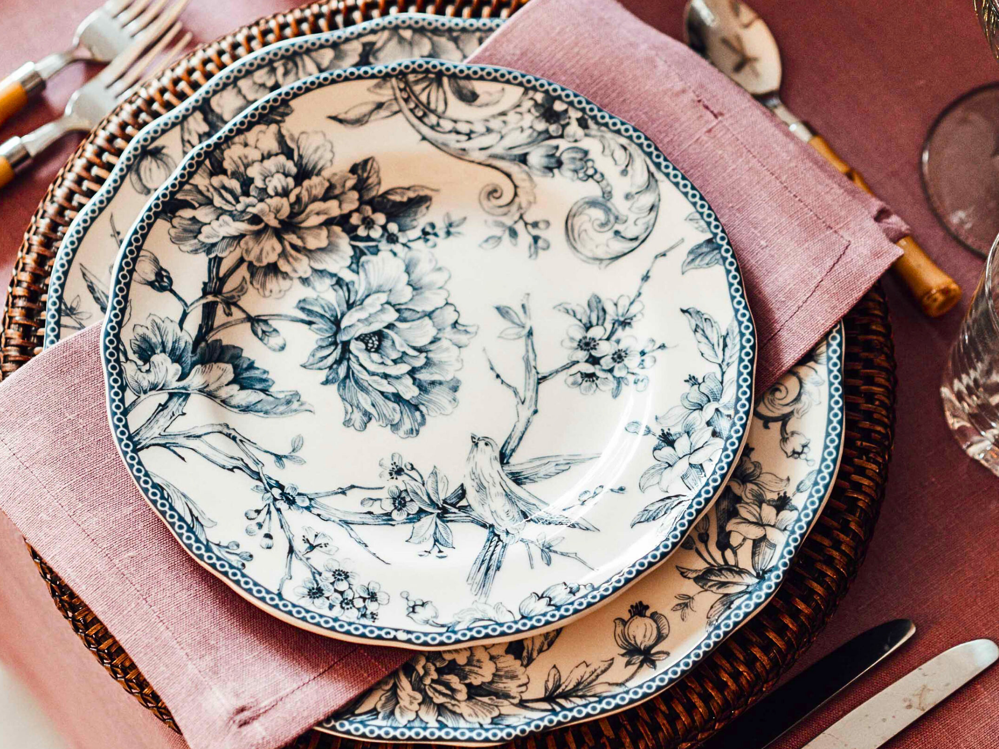 A dining table setting with a pink cloth napkin, blue and white floral patterned plates, clear glasses, arranged on a pink tablecloth.
