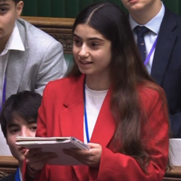 Ealaf stands in Parliament, mid speech, holding a notebook. She wears a red suit and a white shirt. Around her neck is a blue lanyard.