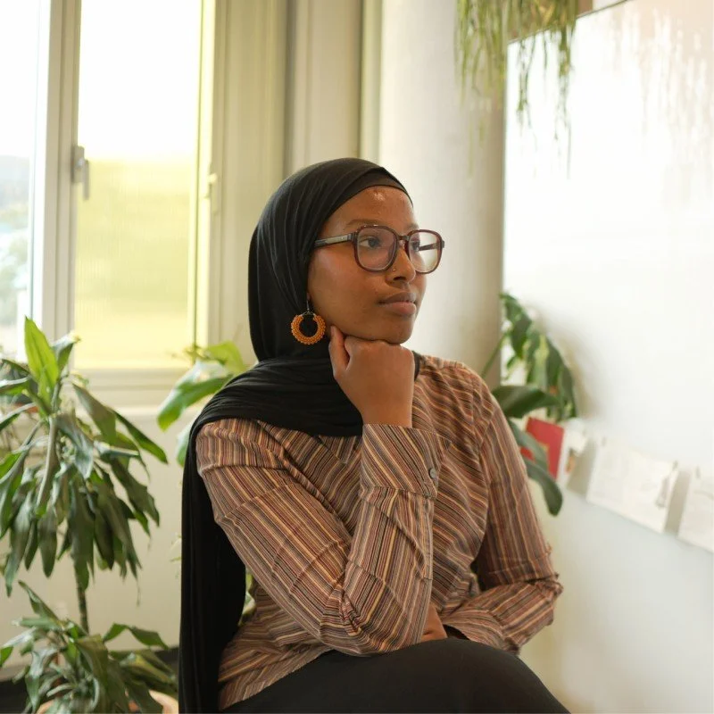An image of Sagal Abdullahi. Sagal is wearing a black headscarf, glasses and thick hooped earrings. She wears a stripy shirt. Her background is light and airy. A plant is just out of focus behind her.