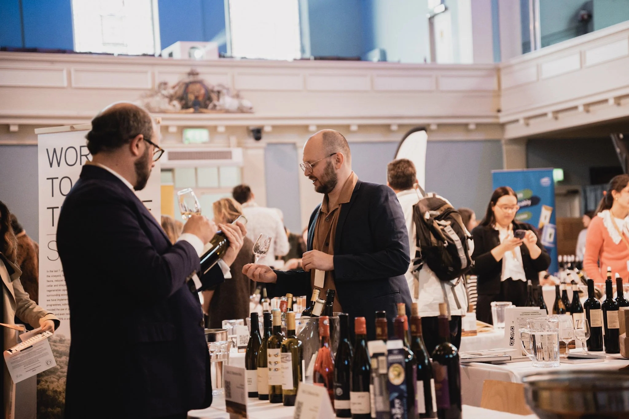 People attending a wine tasting event in a spacious, well-lit hall with wine bottles displayed on tables and a few individuals tasting and discussing wine.
