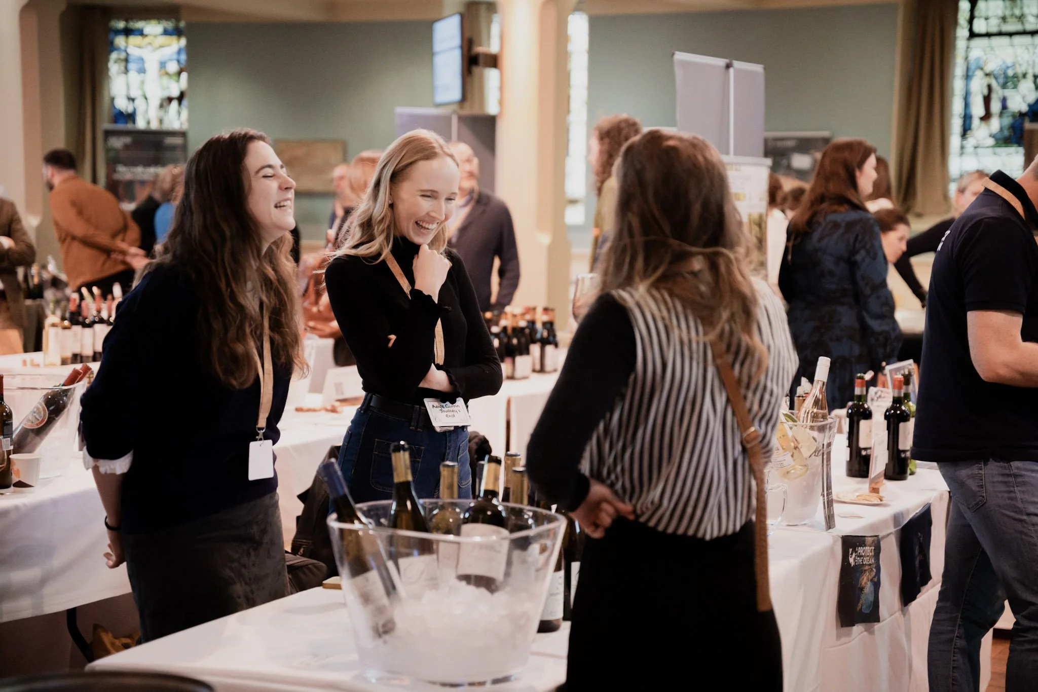 People laughing and chatting at a wine tasting event with tables of wine bottles and a large wine cooler in the foreground.