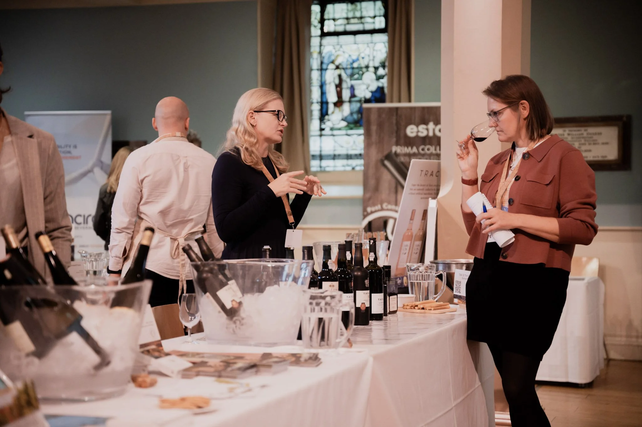 Two women at a wine tasting event, one tasting wine from a glass and the other talking, with bottles of wine and promotional materials on a table.