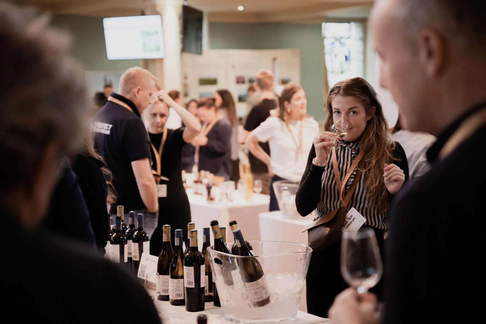 People attending a wine tasting event, with bottles of wine on display and a woman sipping wine, in a bright room with wooden beams.