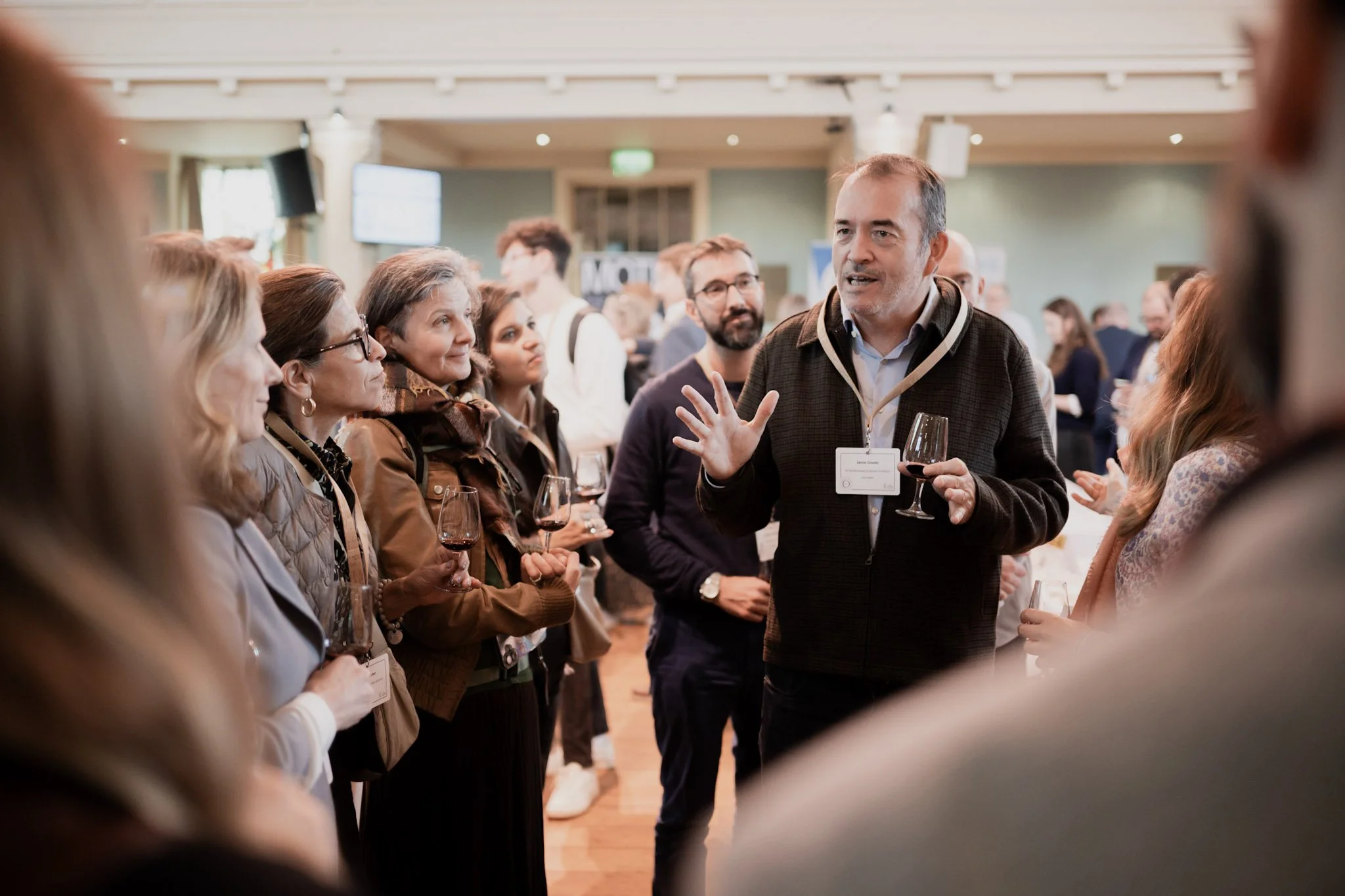 A man in a dark jacket with a conference badge is speaking and gesturing with his hand while holding a glass of red wine, surrounded by people listening at a networking event.