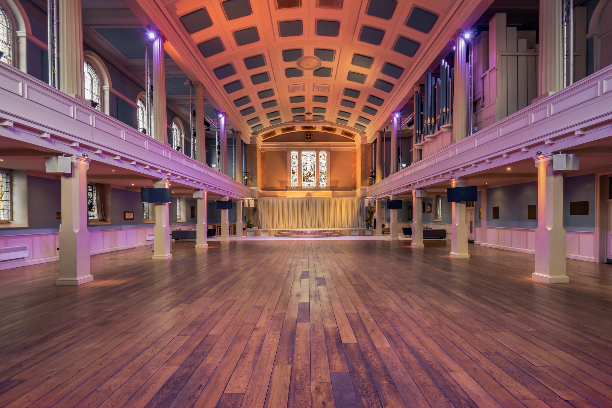 Interior of a spacious hall with wooden flooring, purple and pink lighting, stained glass windows, a stage at the far end, and an ornate ceiling with geometric patterns.